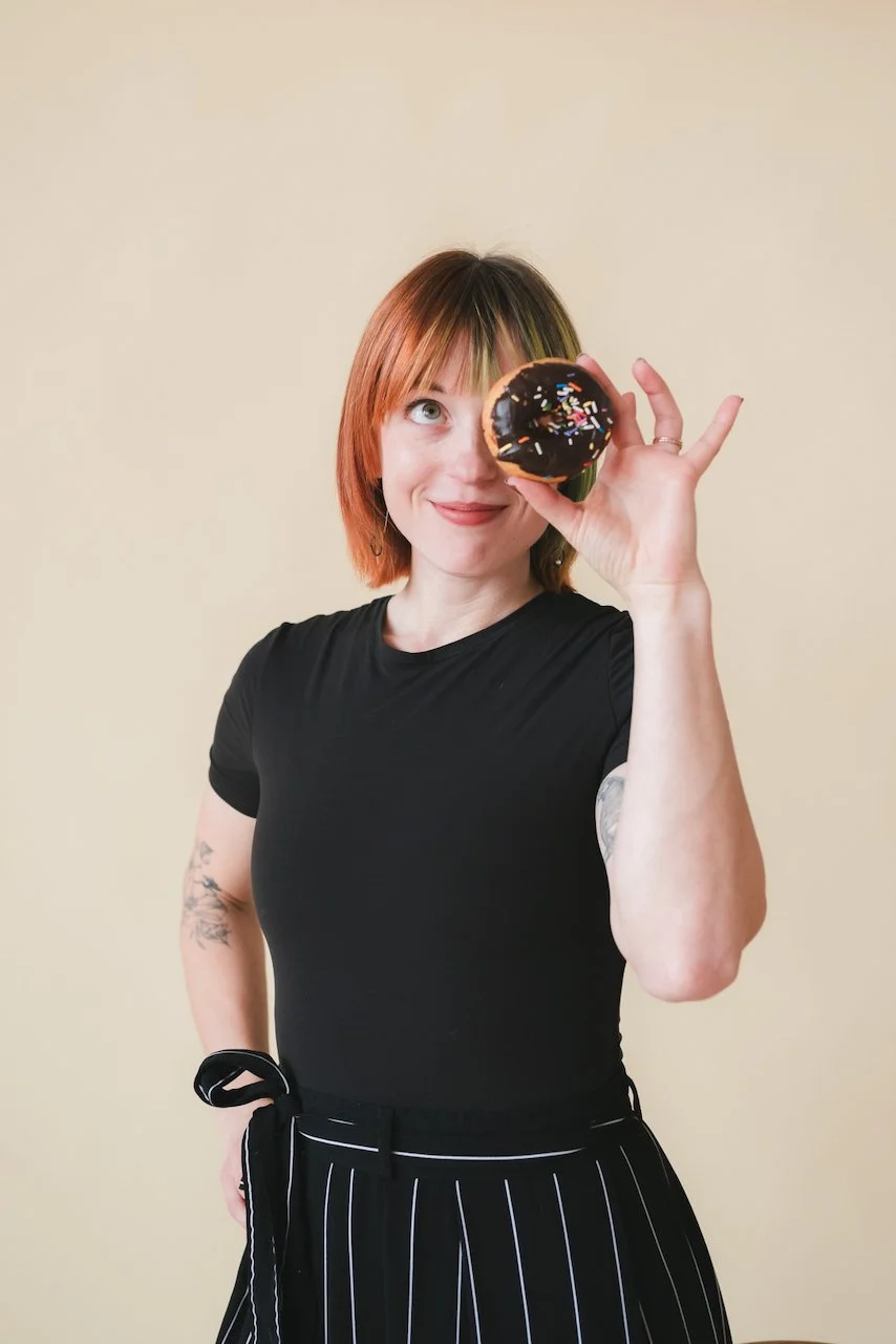 Izy, who is a highly-rated wedding floral designer smiles and holds her favorite post-wedding snack - a chocolate donut, of course - in front of one eye. Image by Kim Nguyen.