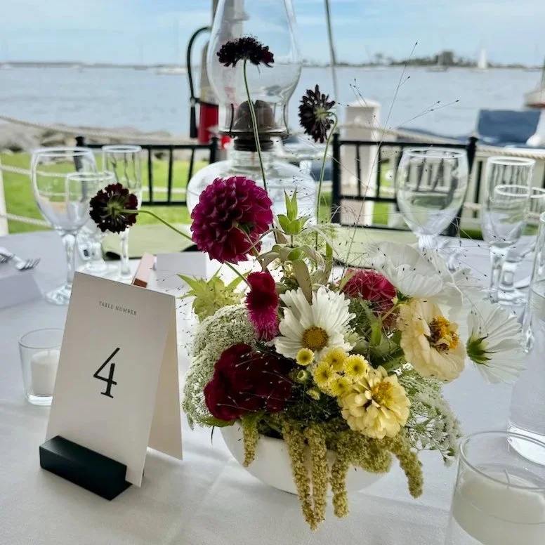 A unique floral centerpiece for a coastal Connecticut wedding on a table set for an event near water, with a table number 4 card. Florals include dramatic shades of plum and chartreuse, with draping green amaranthus.