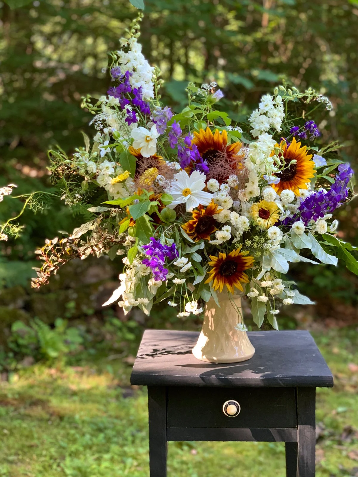 A dramatic and unexpected floral arrangement for a connecticut wedding including sunflowers and seasonal wildflowers in a custom ceramic vase, placed on a small black side table outdoors with green foliage in the background.