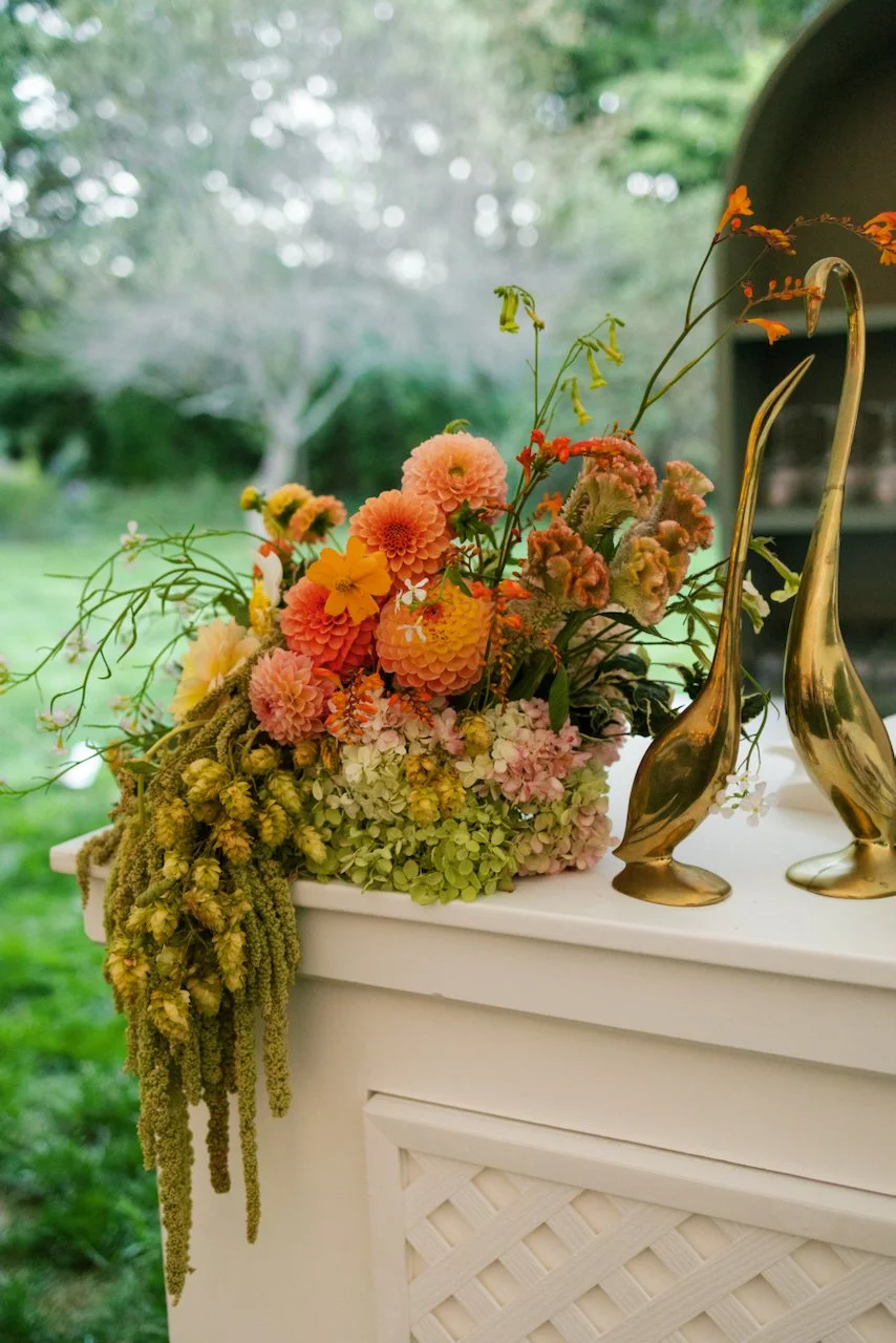 A dramatic floral arrangement with pink, yellow, orange, and white flowers and draping amaranth on a white wedding bar with a garden background, accompanied by two sleek brass animal-shaped figurines.