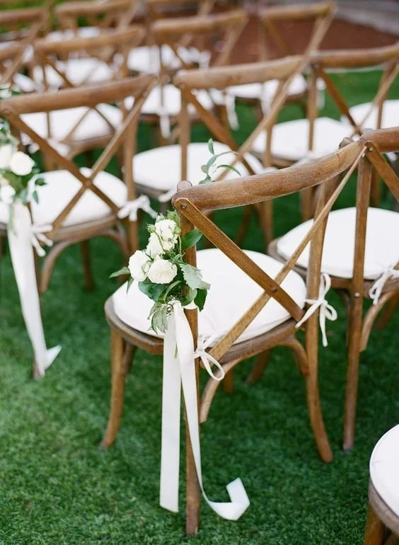 Decorated wooden chairs with white cushions and floral arrangements at an outdoor wedding or event.