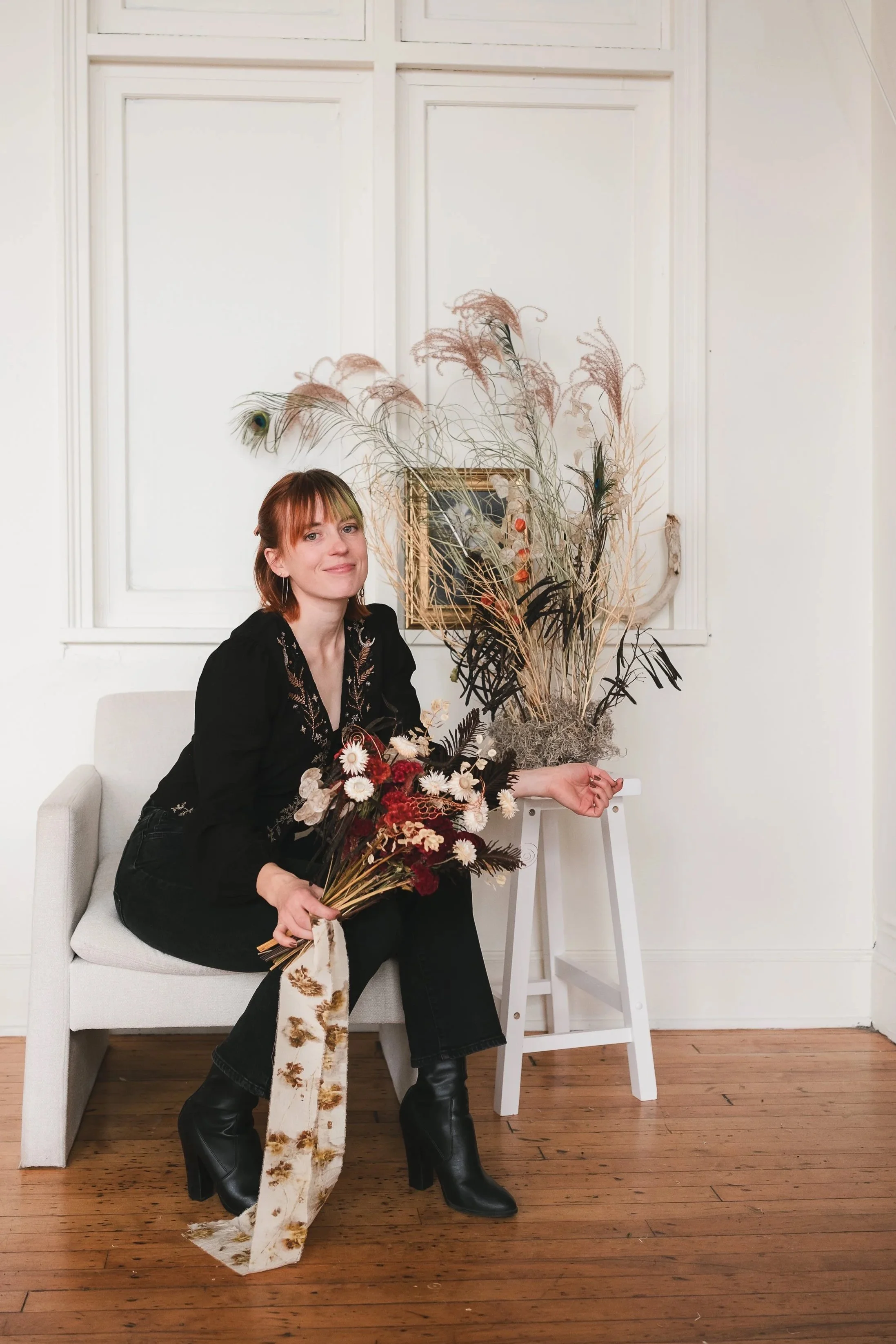 Connecticut Wedding Florist Izy with red hair, wearing black clothing and black high-heeled boots, sitting on a white bench holding a bouquet of vibrant red, soft cream and brown flowers. Behind her is a tall floral arrangement of dried flowers