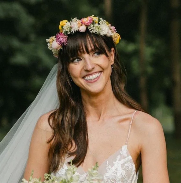 A woman smiling in a wedding dress with a flower crown, outdoors in a forest setting.
