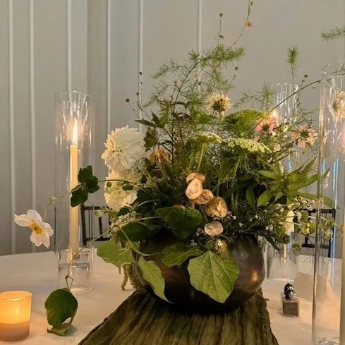 A large unique floral arrangement with dahlias and delicate greenery in a an earthy metal vase, surrounded by candles and glass candleholders on a table.