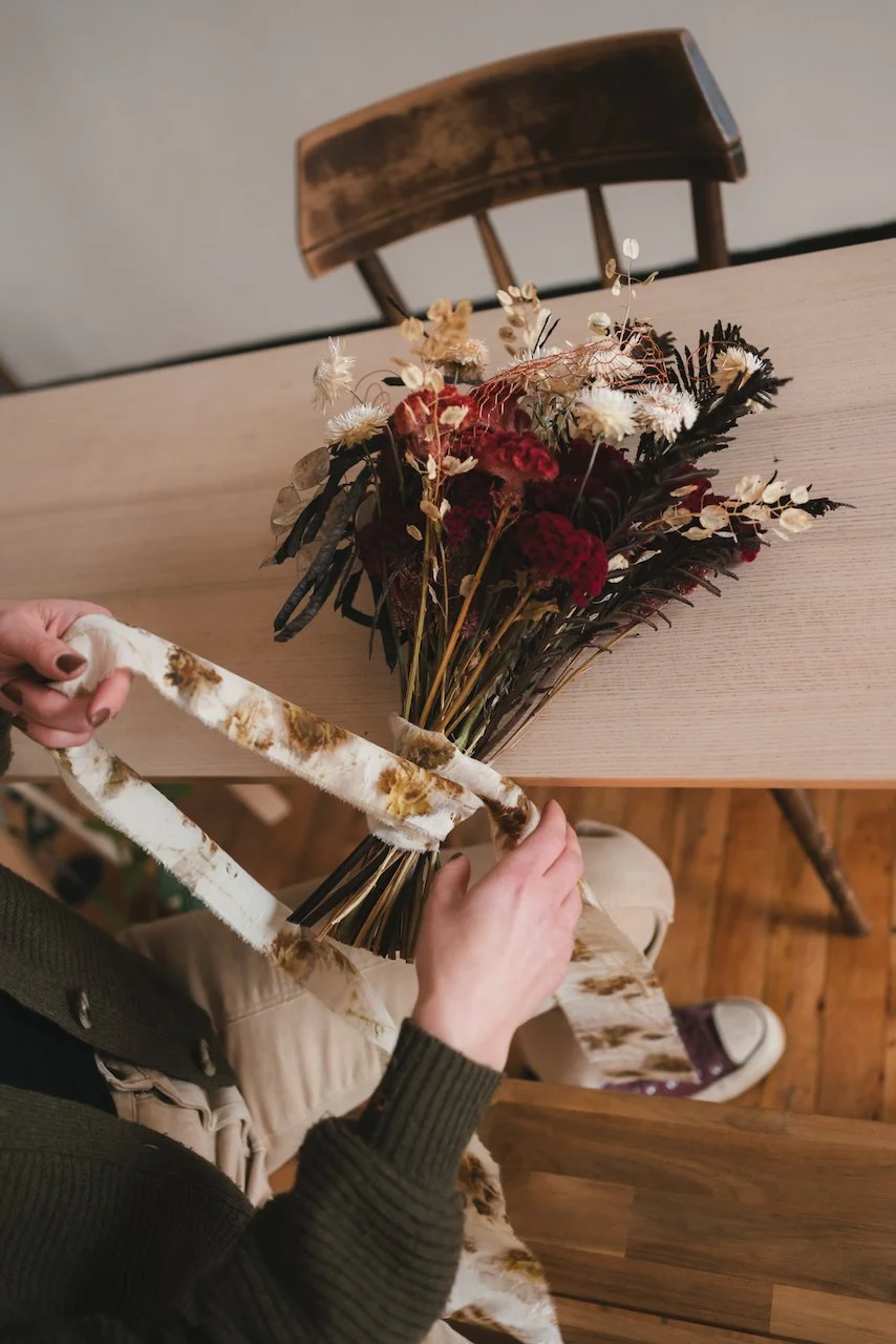 A highly attentive wedding florist sits at a bench and ties a custom silk ribbon onto a rich red, brown, and cream bridal bouquet. The bouquet features unexpected accents of metallic copper wire, woven into interesting shapes. photo by Kim Nguyen
