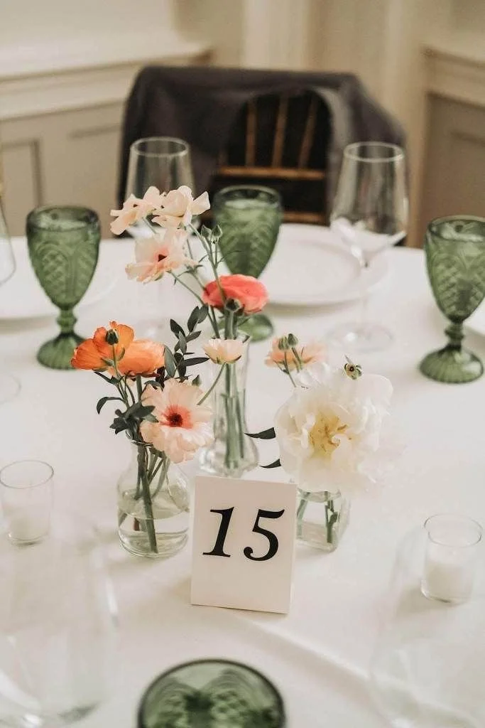 Table setting with floral centerpiece, numbered card 15, green glasses, wine glasses, and small candle holders on a white tablecloth in a formal dining room.