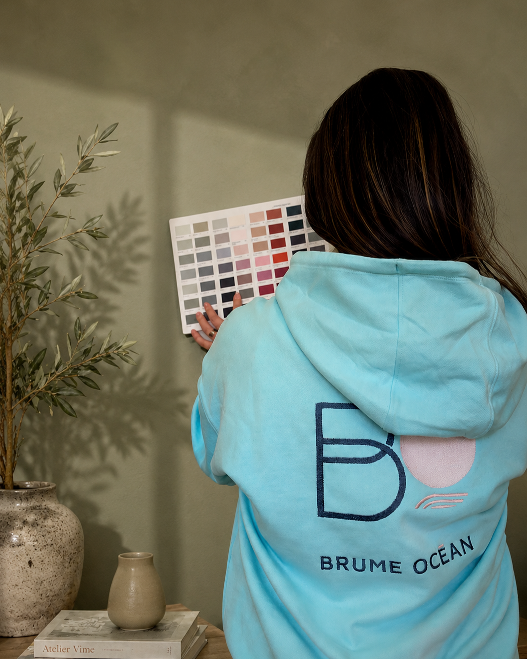 Une femme portant un sweat à capuche bleu regarde un échantillonneur de couleurs sur un mur.
