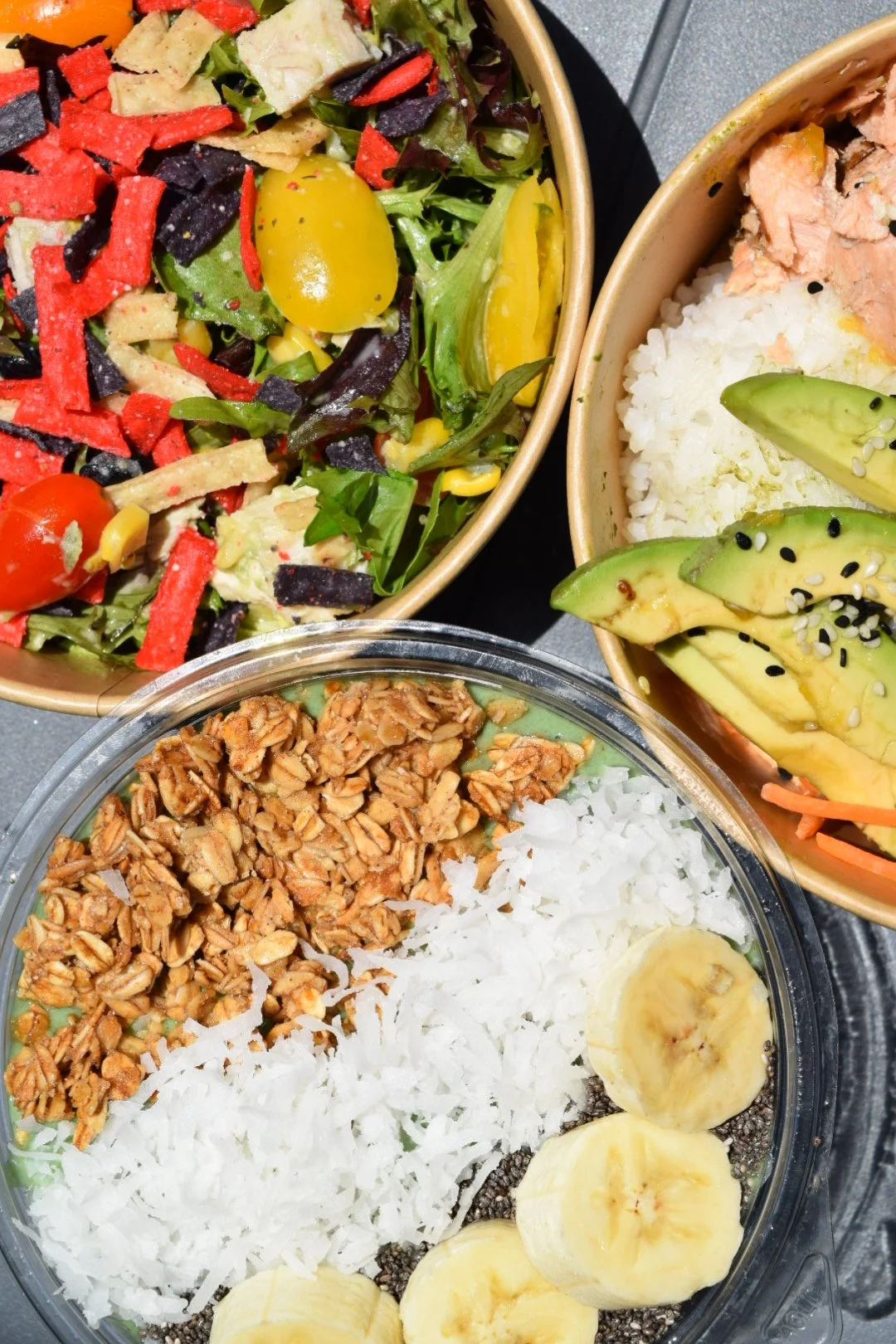 a bird's eye view of three food bowls: a salad bowl with colorful tortilla strips and tomatoes, acai bowl topped with granola, coconut, chia, and banana, and a rice bowl with avaocado and salmon.