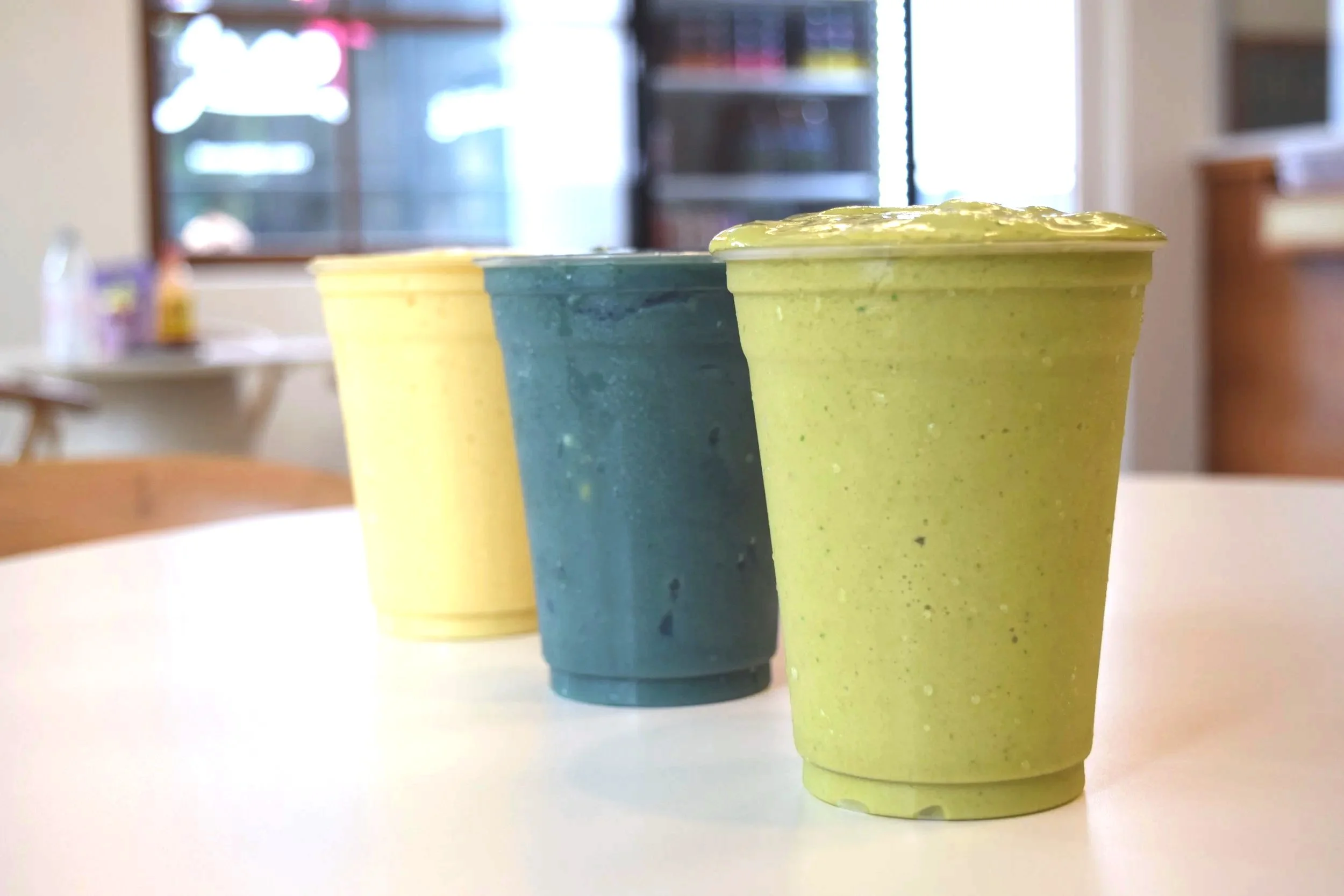 Three smoothies in plastic cups with lids, displayed in a row on a white surface, in front of a blurred background of a room with shelves and a window.