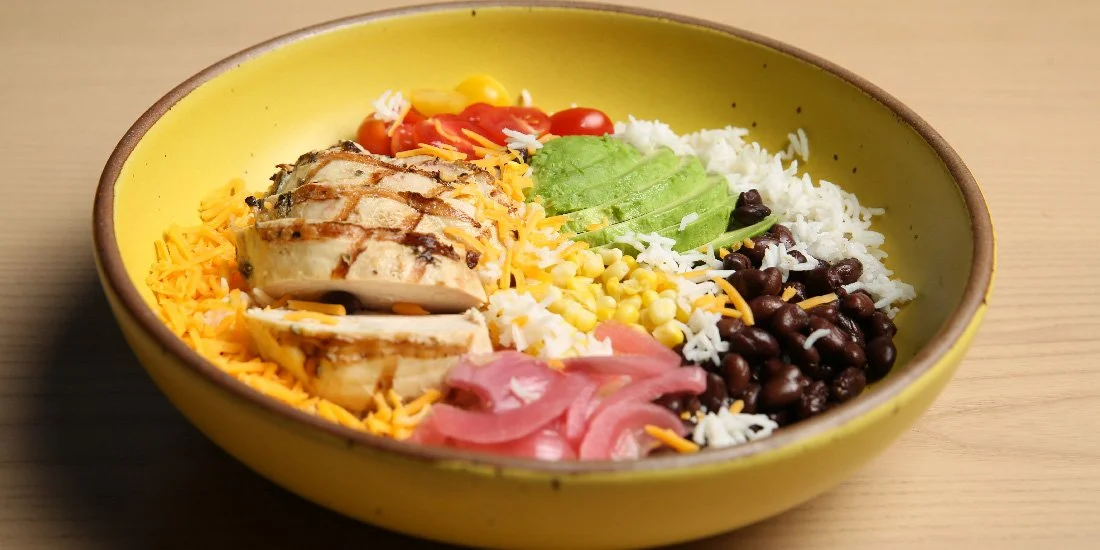 A yellow bowl filled with salad ingredients including grilled chicken slices, shredded cheese, cherry tomatoes, sliced avocado, black beans, corn, shredded cheese, and pickled onions.