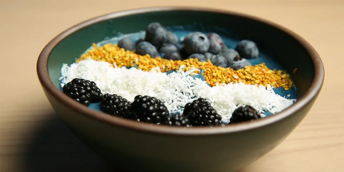 A bowl with blueberries, blackberries, shredded coconut, bee pollen, and granola on top of a blue smoothie.