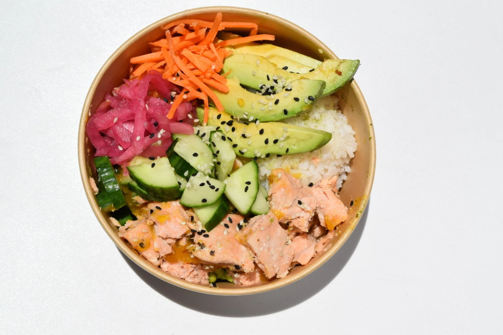 a paper bowl containing white rice, salmon, avocado, cucumber, pickled red onions, shredded carrots, and sesame seeds.
