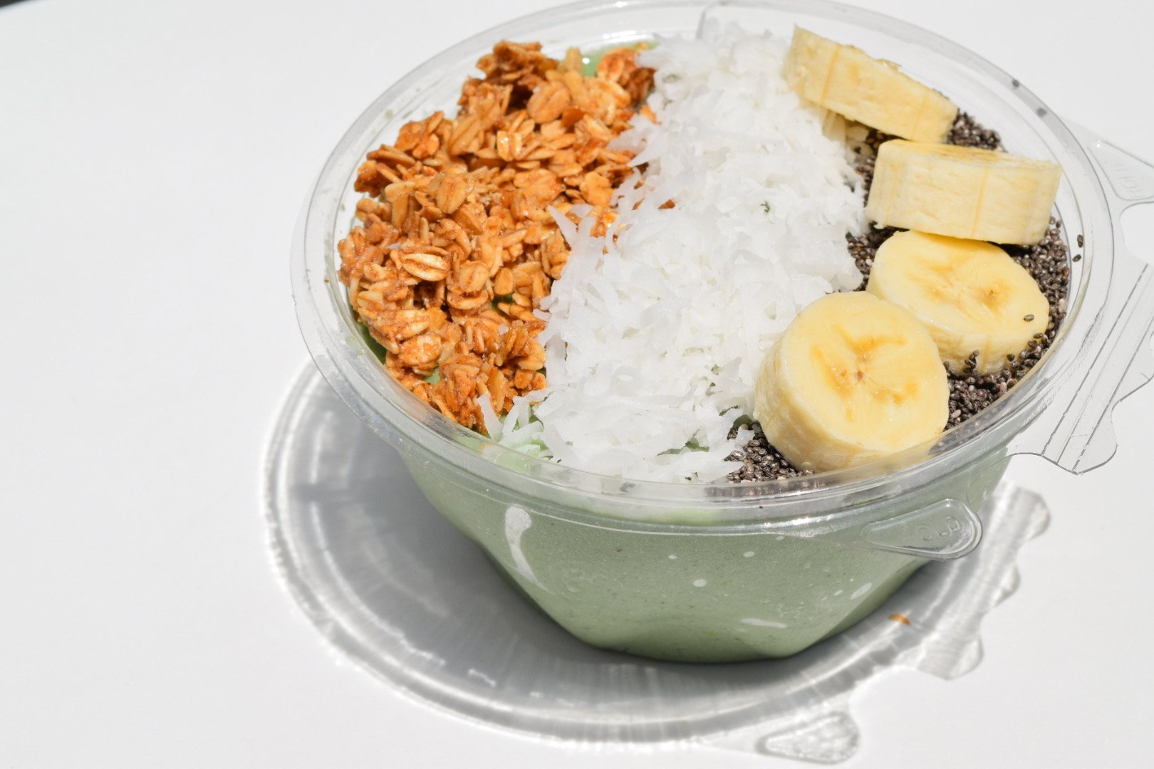 green acai bowl in a plastic container topped with granola, coconut, banana and chia seeds