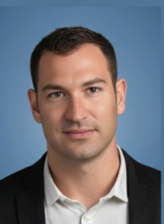 A man in a dark suit and white shirt standing outdoors at night with blurred city lights in the background.