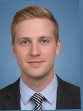 A smiling man with reddish hair, wearing a light gray dress shirt and a black and white striped tie, standing outdoors at night with blurred colorful lights in the background.