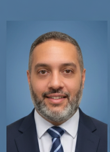 A man with short, dark hair and a beard, wearing a dark suit and a striped tie, standing outdoors at night with blurred city lights in the background.