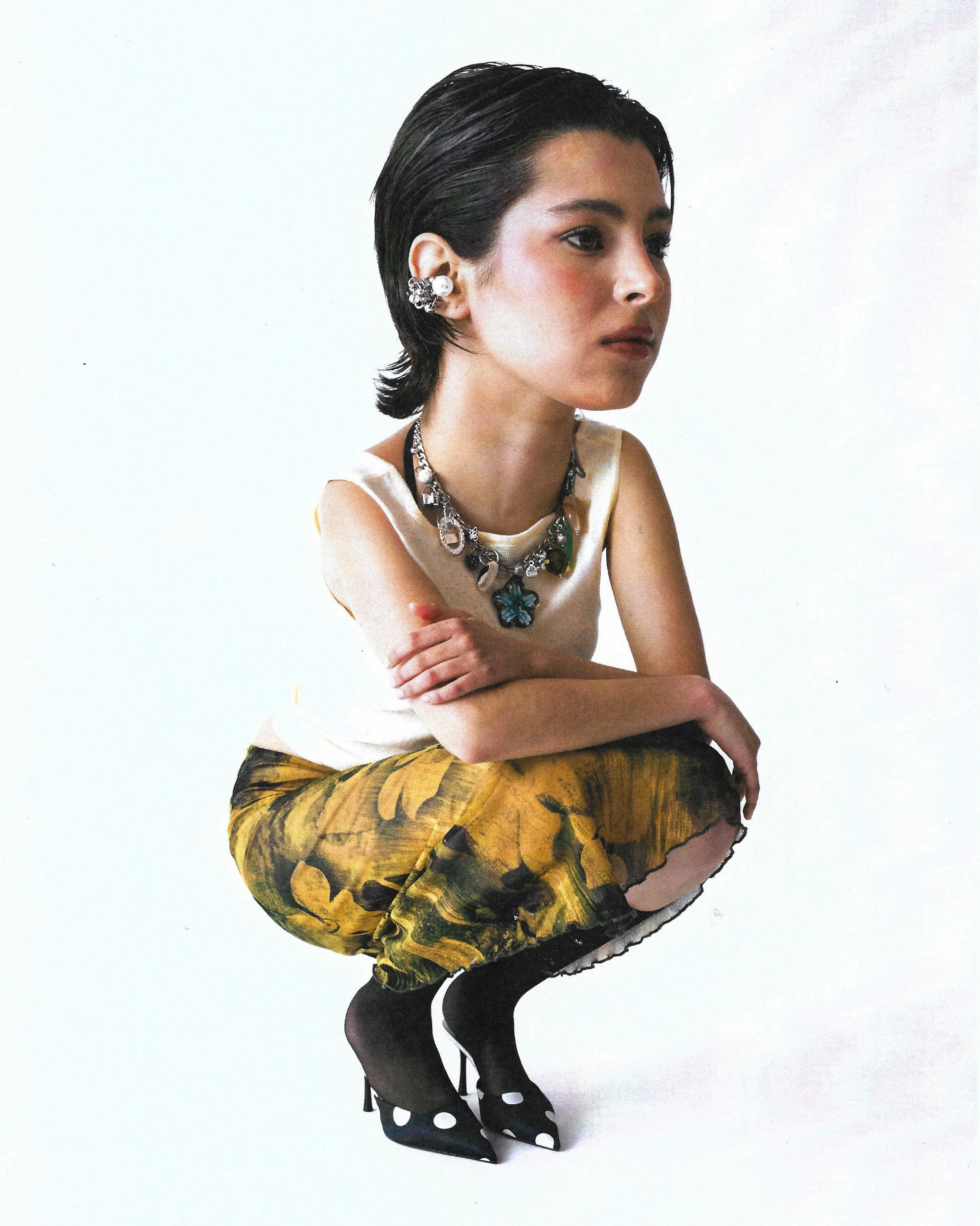 A woman with dark, short hair, crouching with arms crossed over her knees against a plain white background. She is wearing a beige sleeveless top, a large, colorful necklace, decorative earrings, a yellow and black patterned skirt, black tights, and 