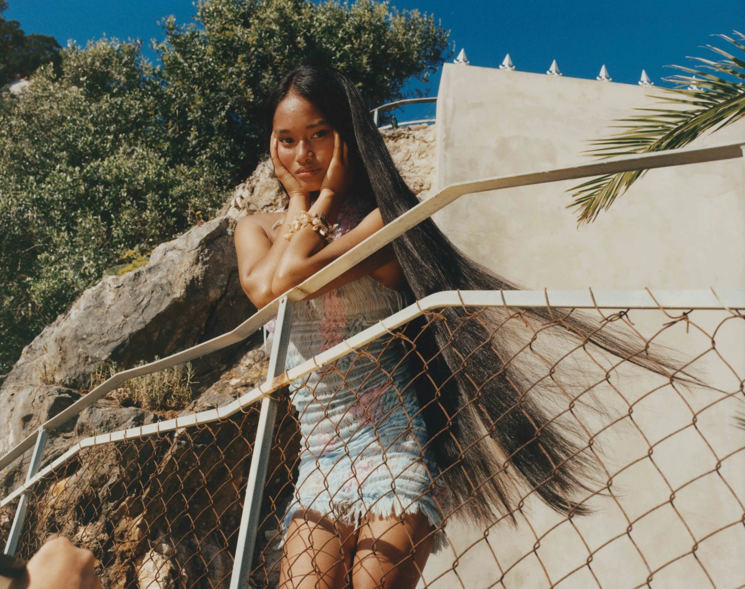 Young girl with long dark hair leaning on a railing, looking at the camera, wearing a sleeveless top and shorts, outdoors with rocks and greenery behind her, sunny day with clear blue sky.