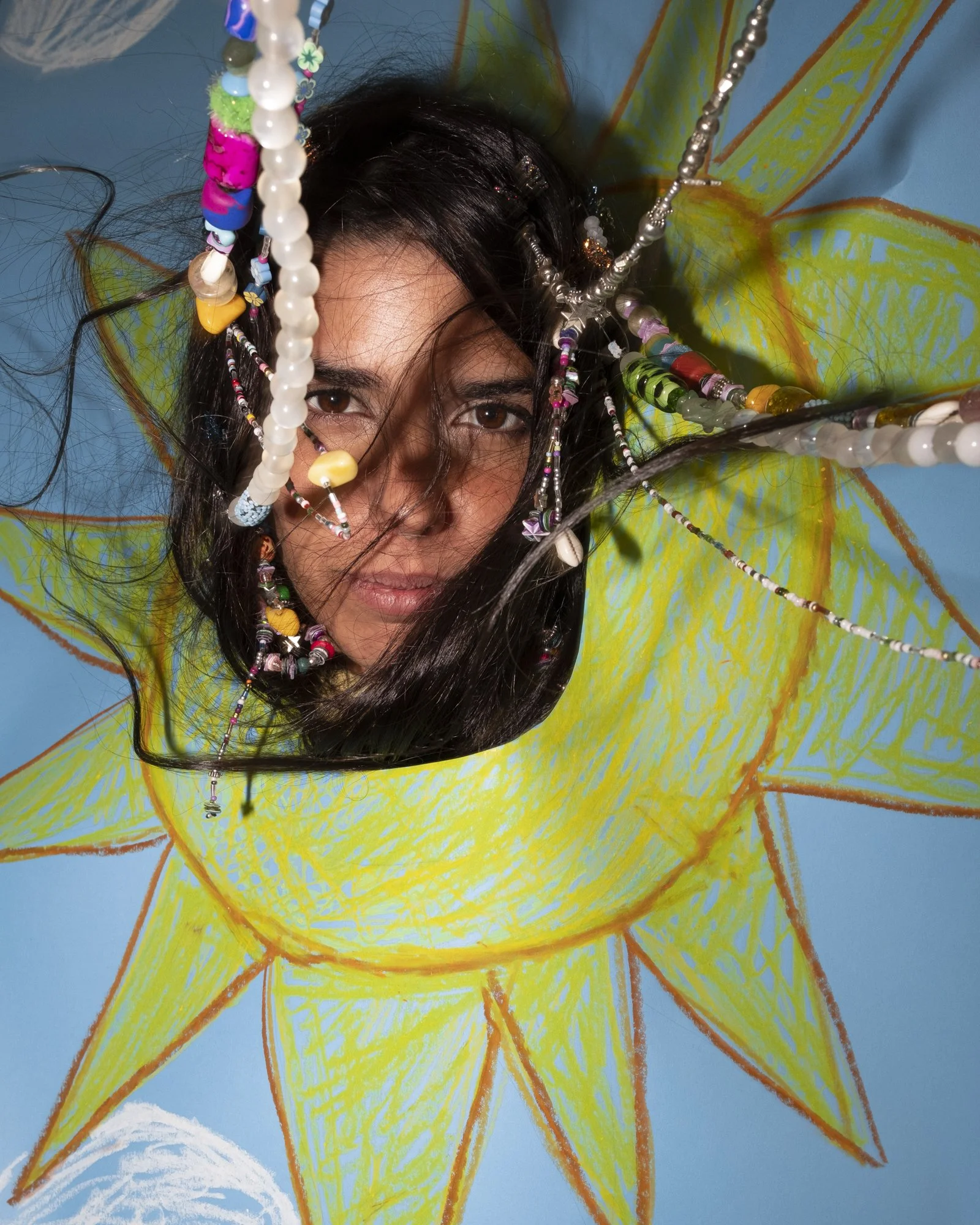 A woman with long dark hair and brown eyes looking directly at the camera through a hole in a painted blue background with a large yellow sun design. She has various colorful beaded necklaces draped across her face and head.