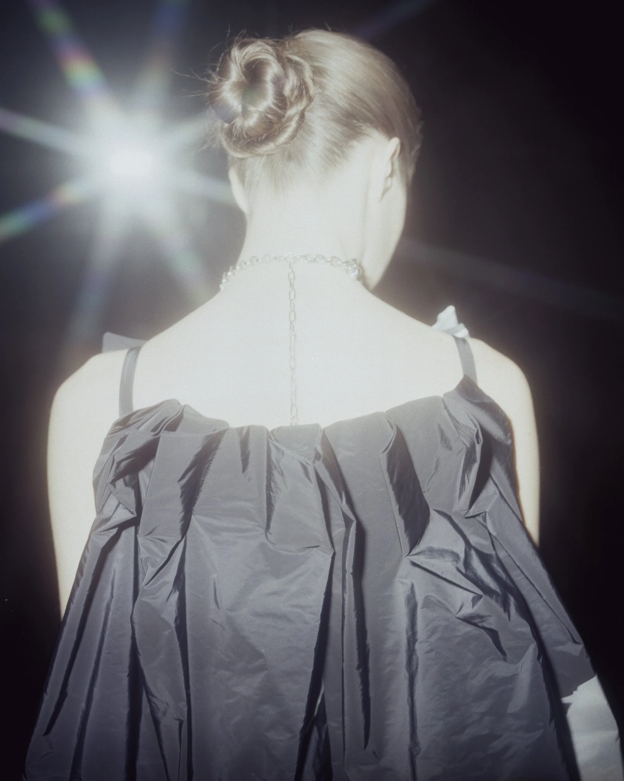 Back view of a woman with styled hair, wearing a dark dress with ruffled sleeves, and a chain necklace, illuminated by a bright light source from the back.