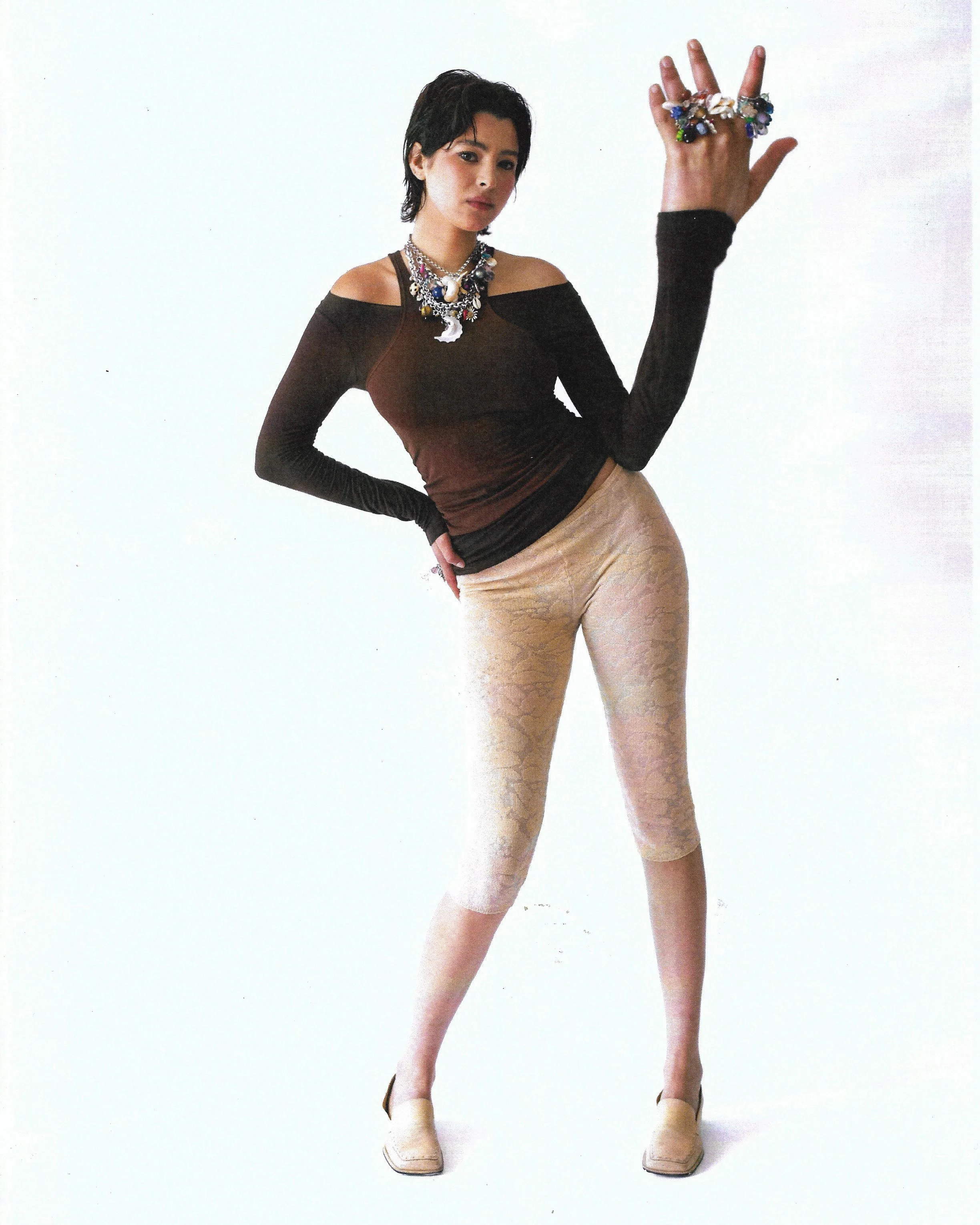 A woman with short black hair wearing a black off-shoulder top, beige patterned leggings, beige shoes, and multiple colorful jewelry pieces, standing with one hand on her hip and the other raised, against a plain white background.