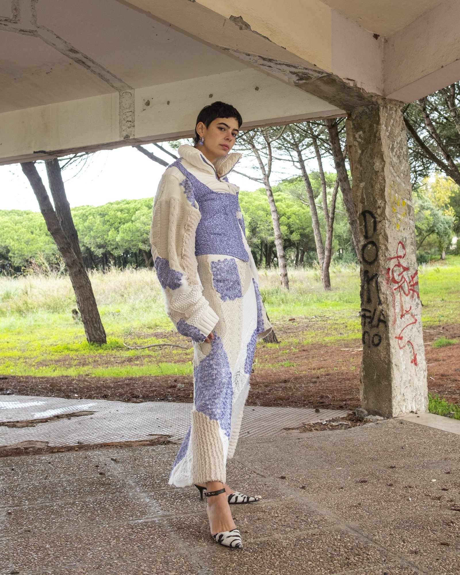 A woman standing outdoors under a concrete structure, wearing a patchwork knitted outfit with purple and cream colors, high heels, and earrings, with a background of green trees and graffiti on a concrete pillar.