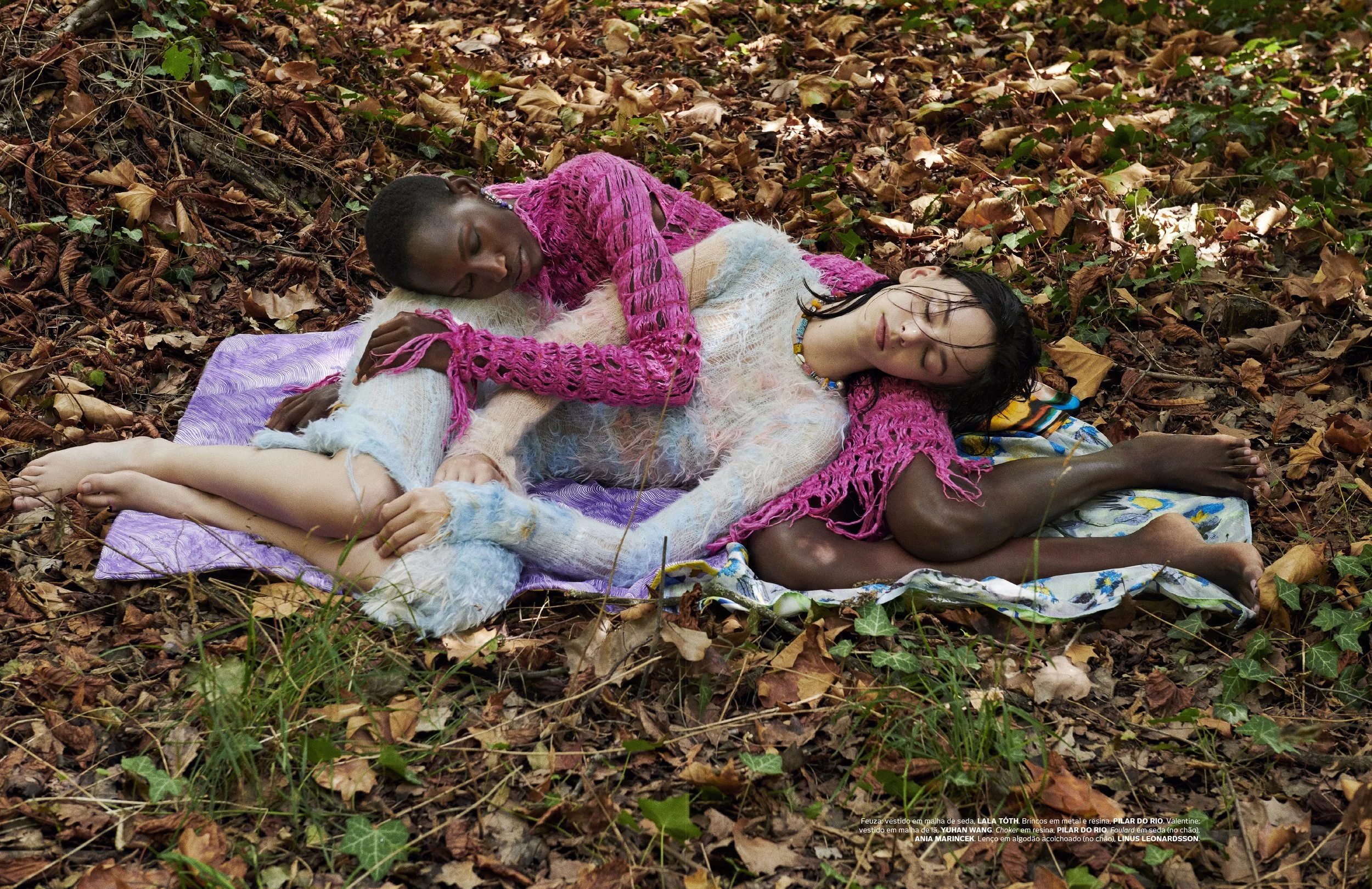 Two women lying on a blanket in a wooded area with fallen leaves, one with dark skin and the other with light skin, both wearing colorful knitwear and jewelry, appearing to rest or nap.