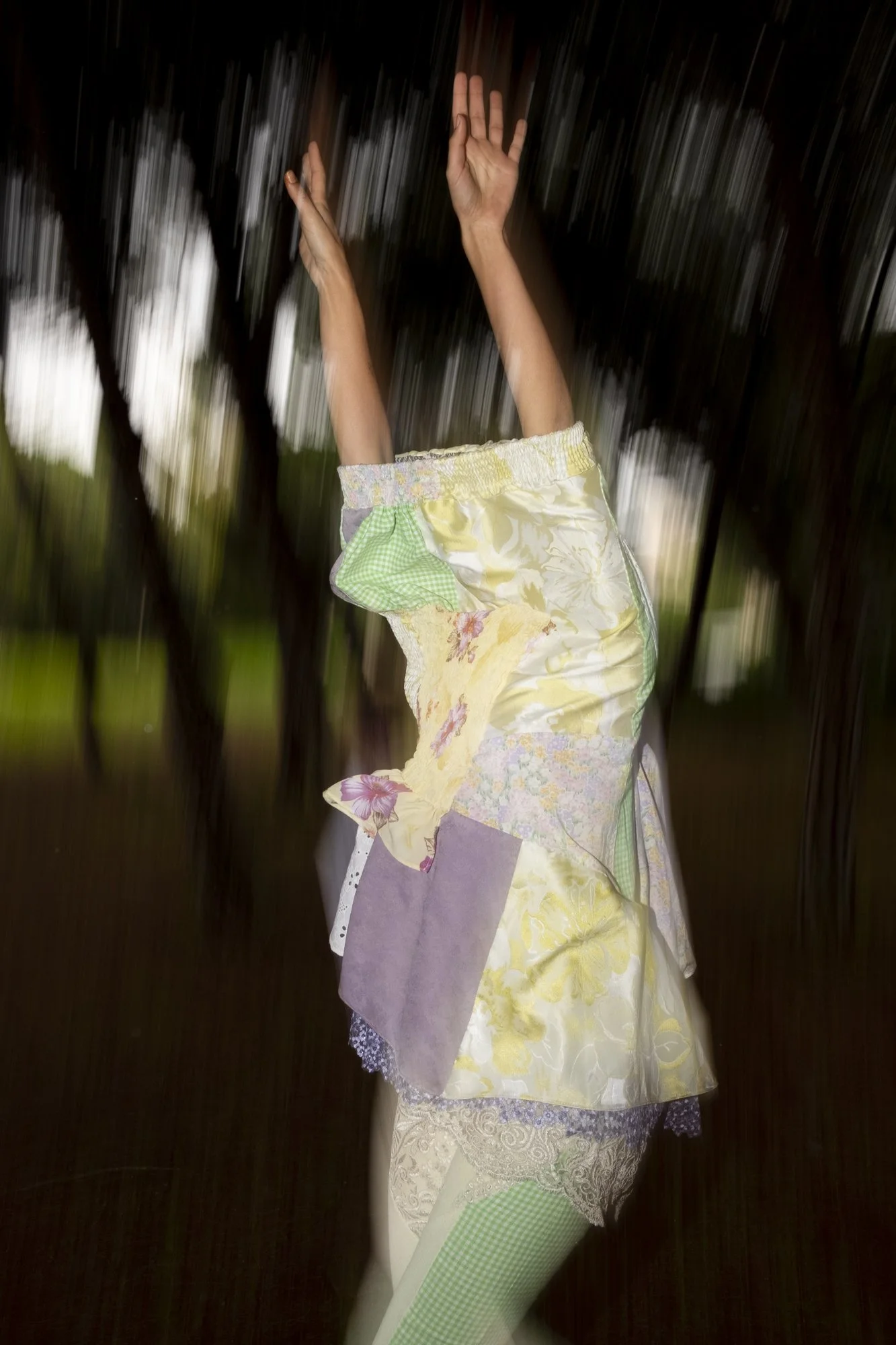 A person is running  with arms raised and head near the ground, wearing a colorful patchwork dress and leggings, outdoors with blurred trees in the background.