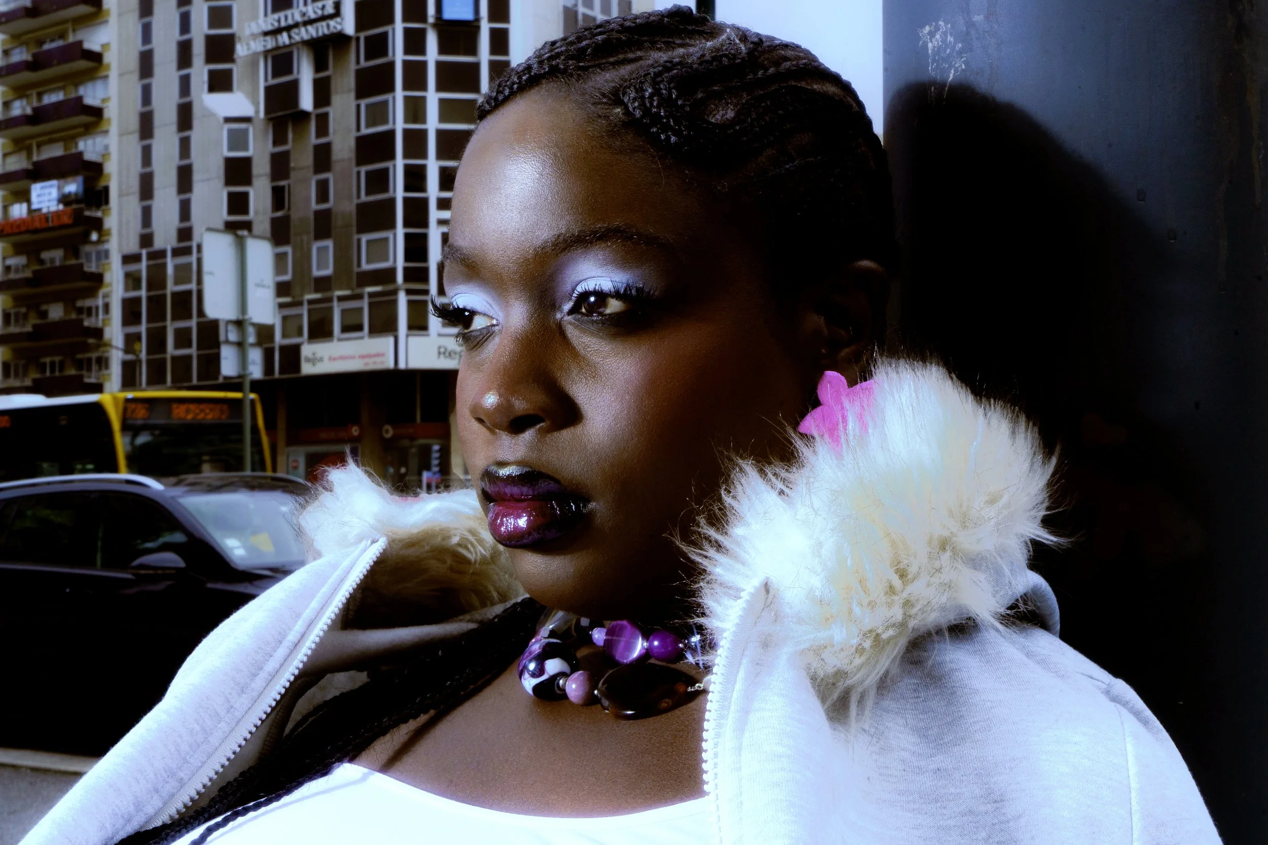 A woman with braided hair, purple lipstick, and a purple necklace stands outdoors in an urban setting with tall buildings, parked cars, and a bus in the background.