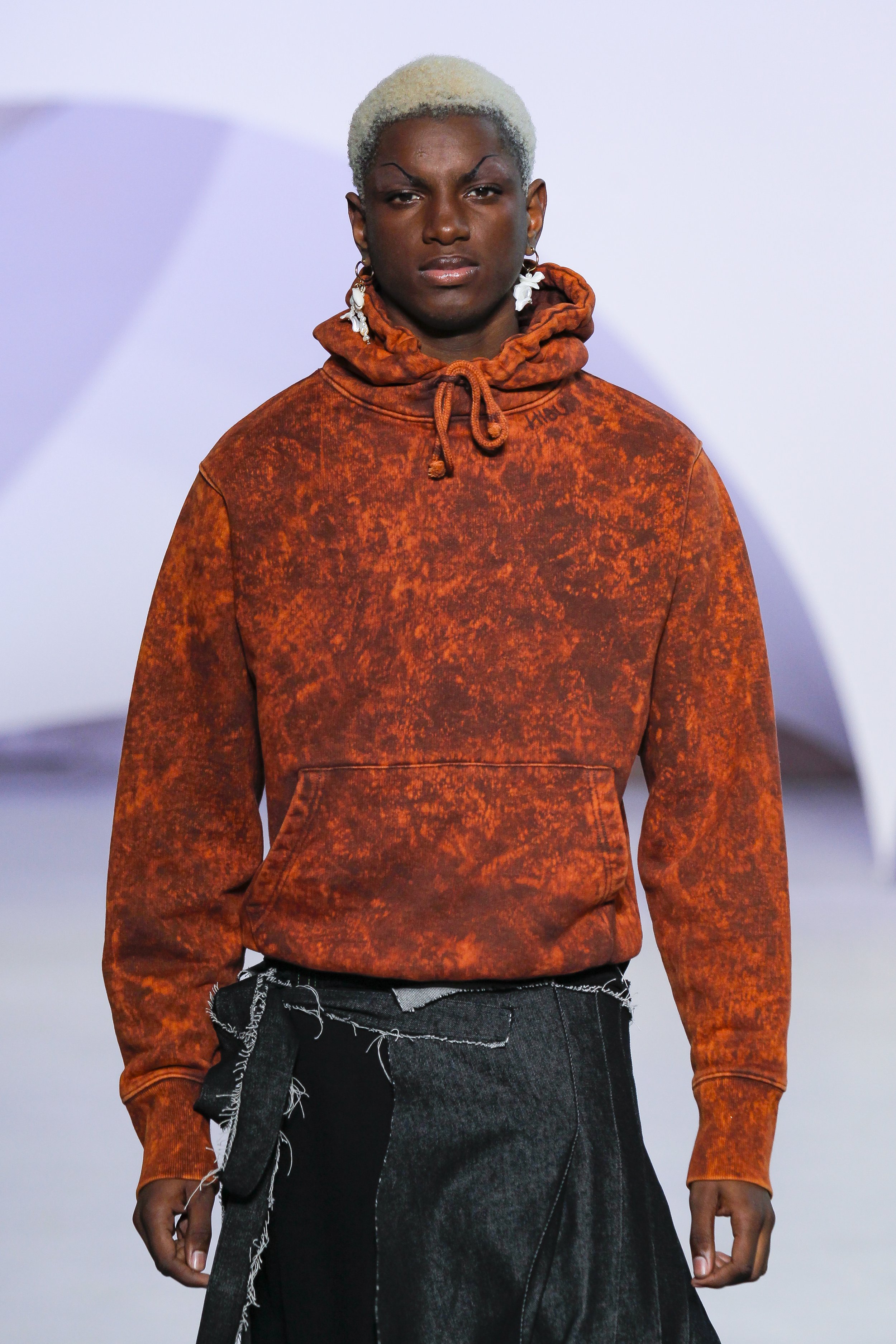 Fashion model wearing an orange, distressed hoodie and black skirt with frayed edges on a runway.