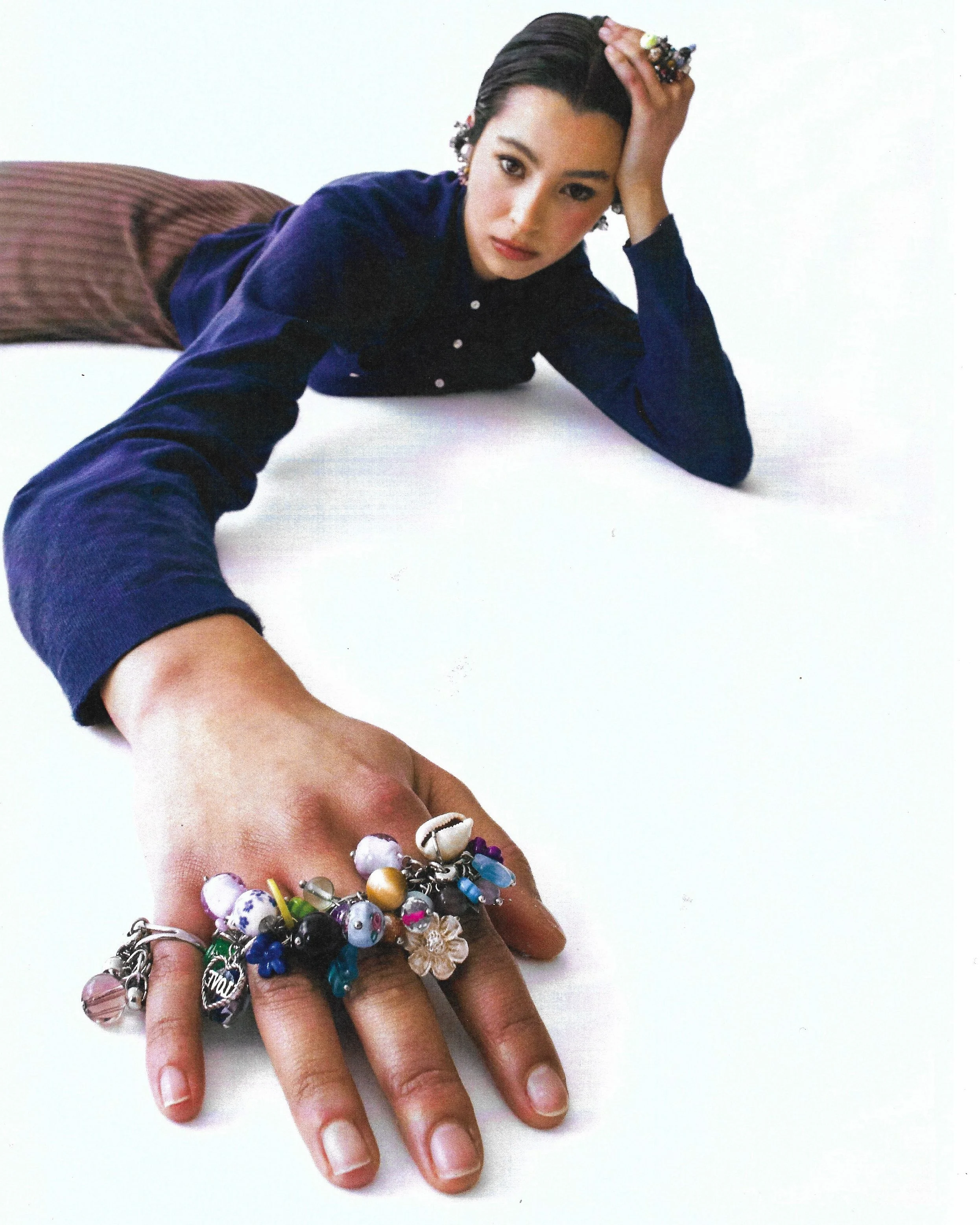A woman lying on her stomach with her arm extended towards the camera, showcasing a collection of colorful bracelets on her fingers, wearing a navy blue shirt and brown pants.
