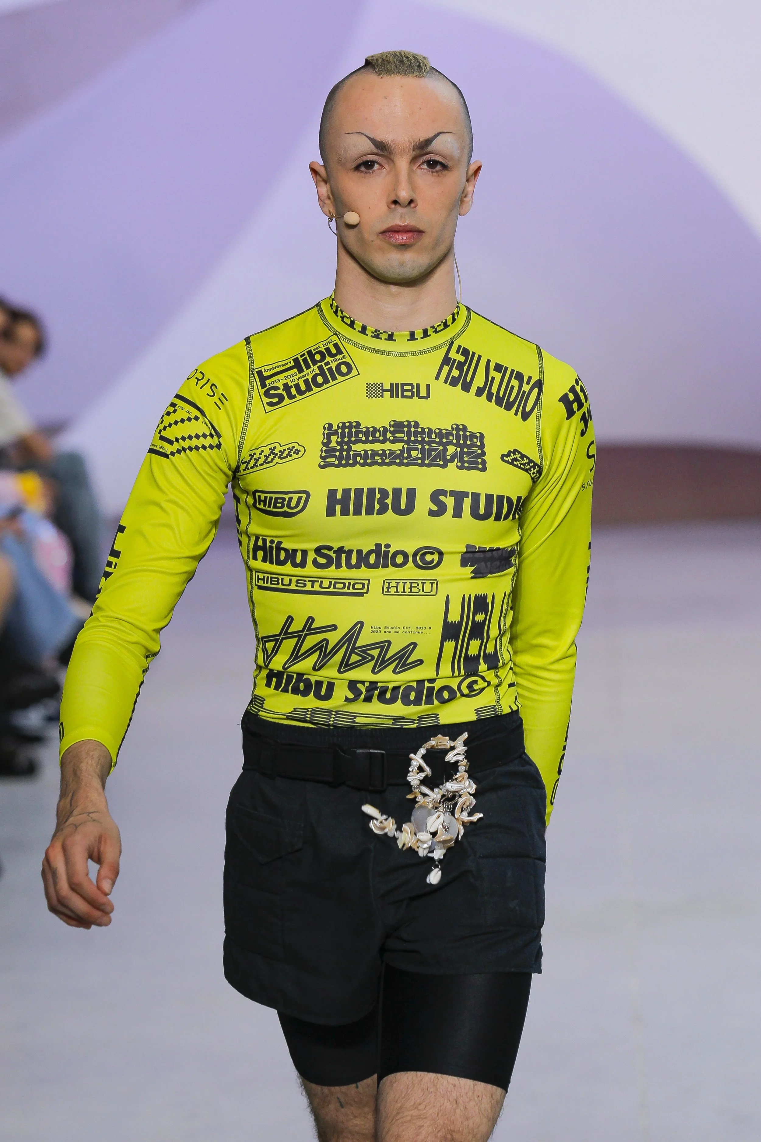A male model walking down a runway during a fashion show, wearing a bright green long-sleeve shirt with black text and logos, black shorts with gothic jewelry, and a microphone on his face.