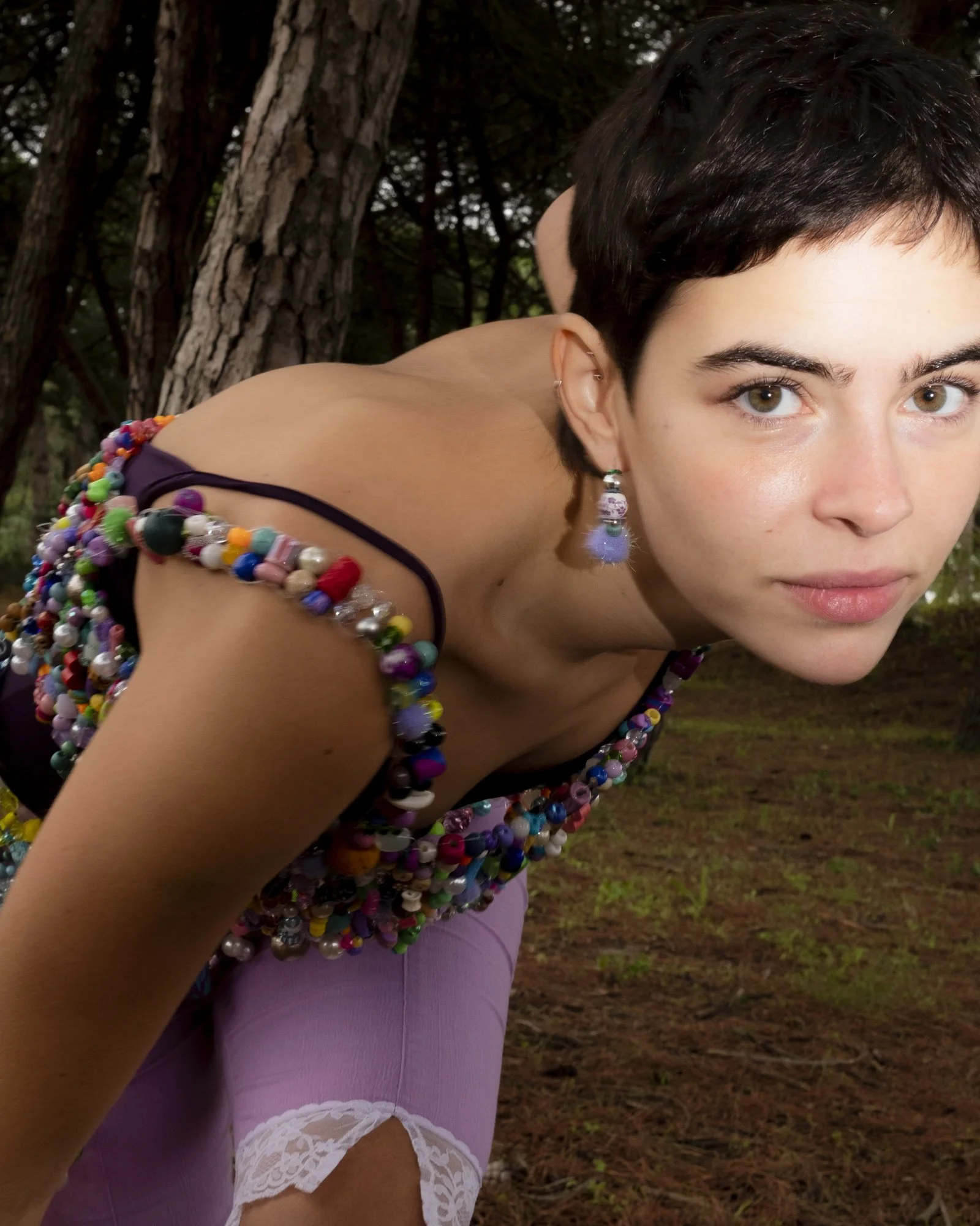 A young woman with short dark hair and earrings, wearing a colorful beaded top and pink pants with lace details, poses outdoors near trees.