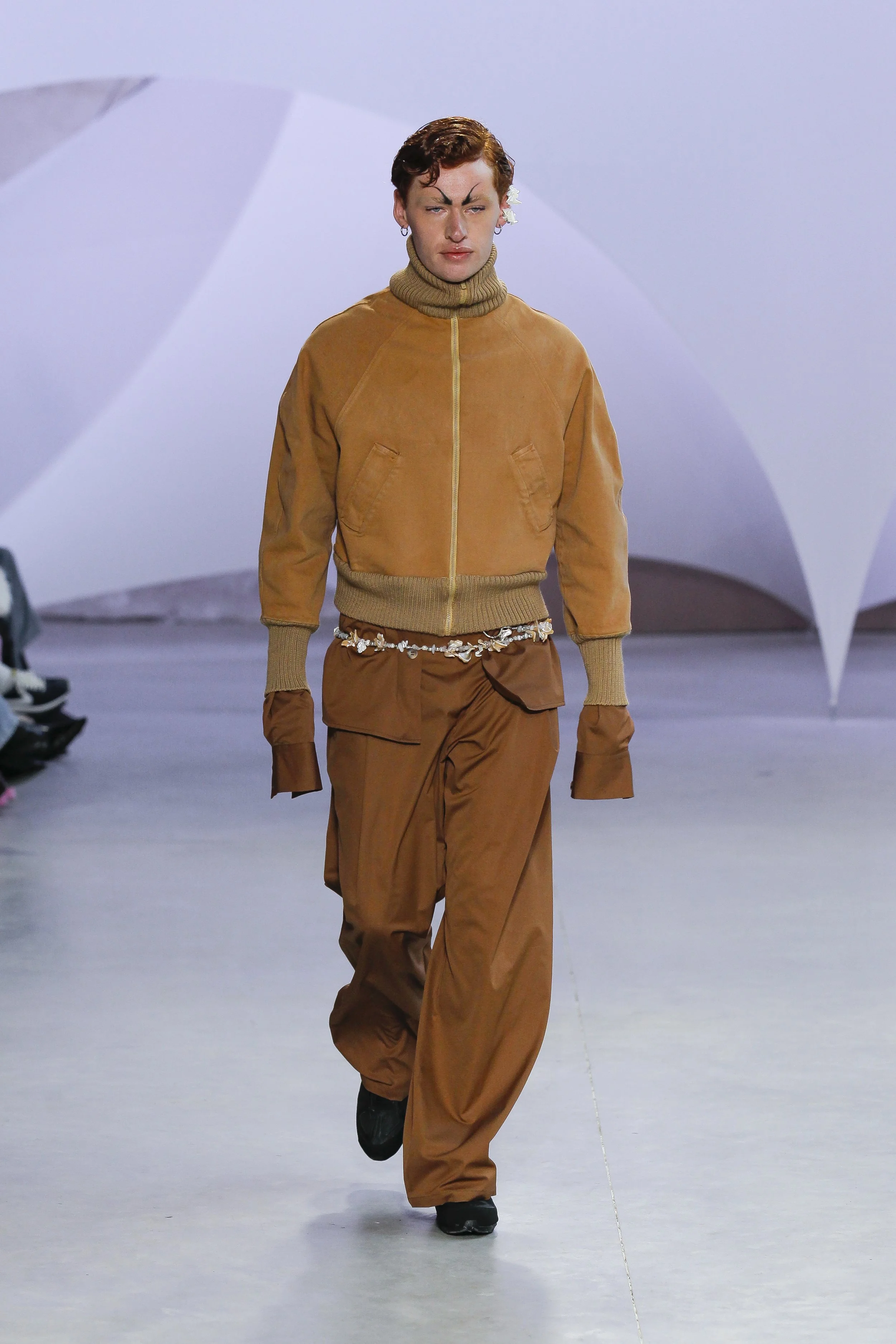 A male model walking on a runway in a fashion show, wearing a tan jacket, matching loose pants, and a decorative belt. The background features white geometric structures.