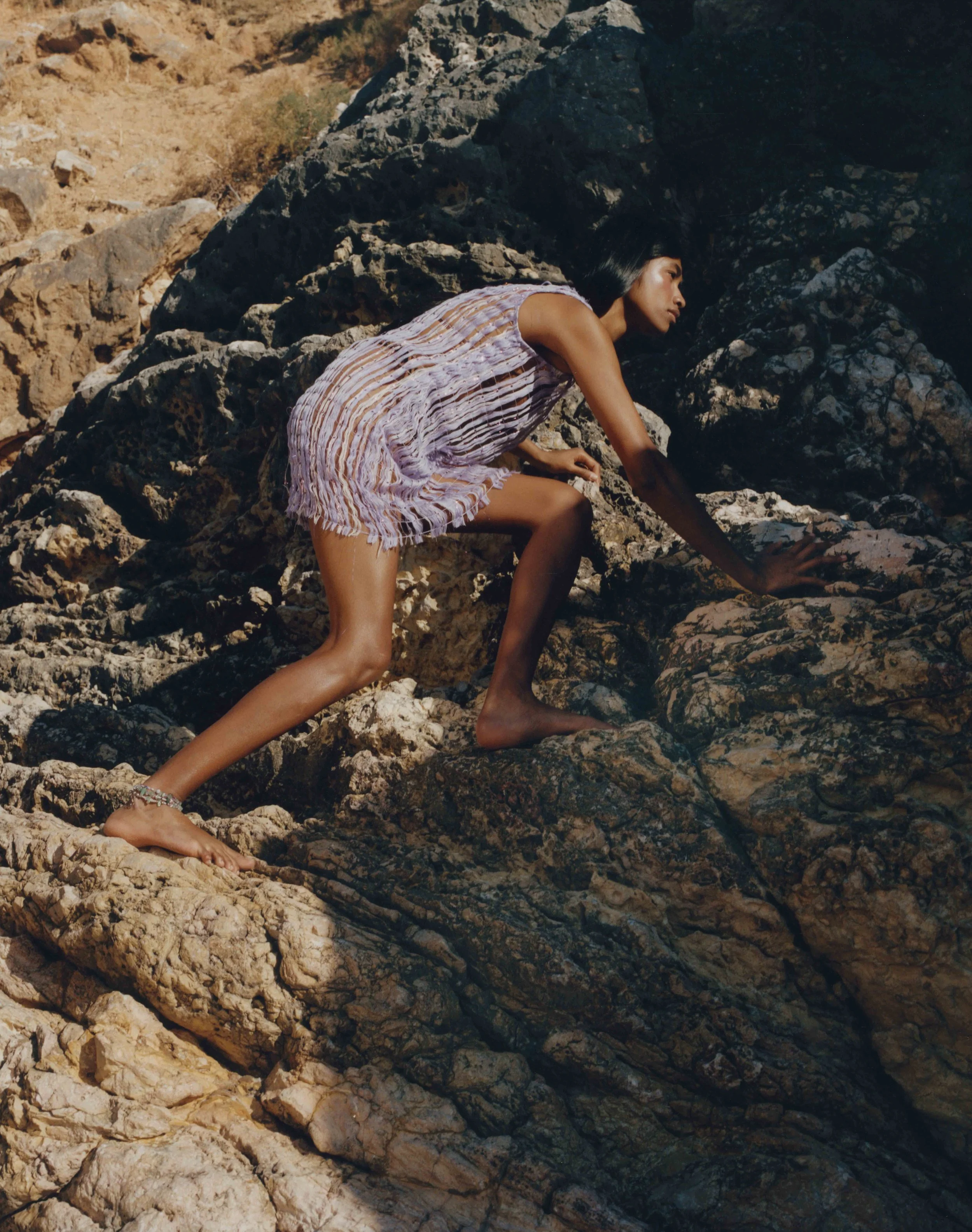 A woman climbing rocky terrain outdoors during daytime, wearing a striped, fringed dress.