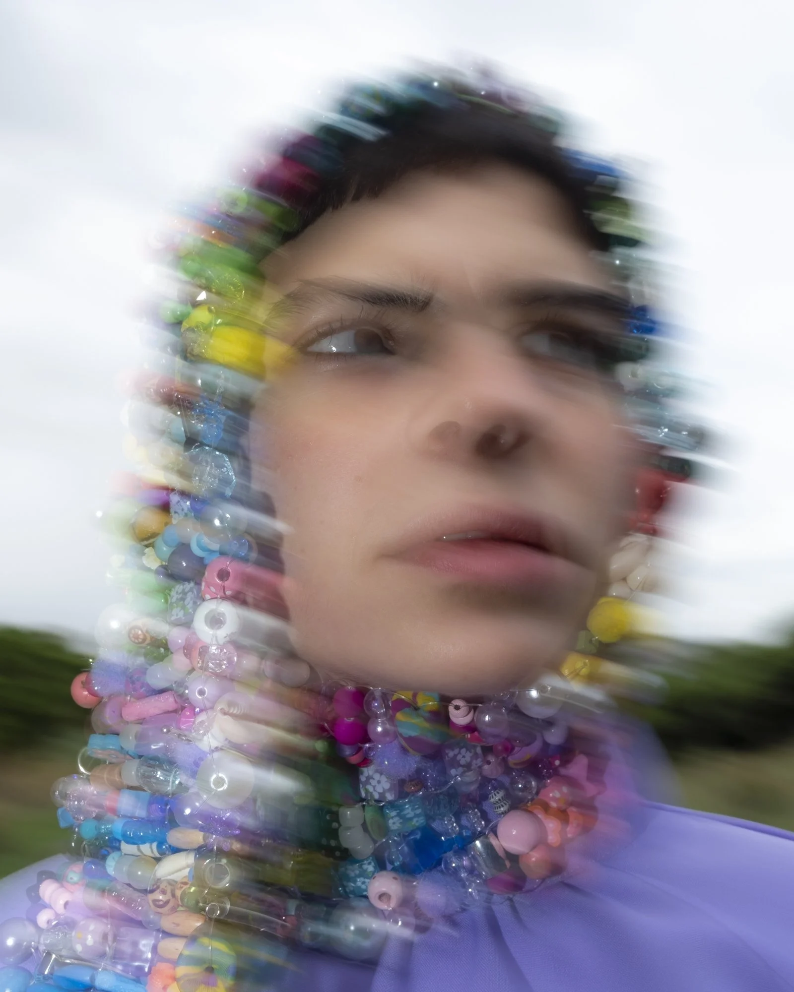 A person with short dark hair, wearing a beaded necklace, is outdoors with a blurred background. The image is intentionally blurred around the face, creating a motion effect.