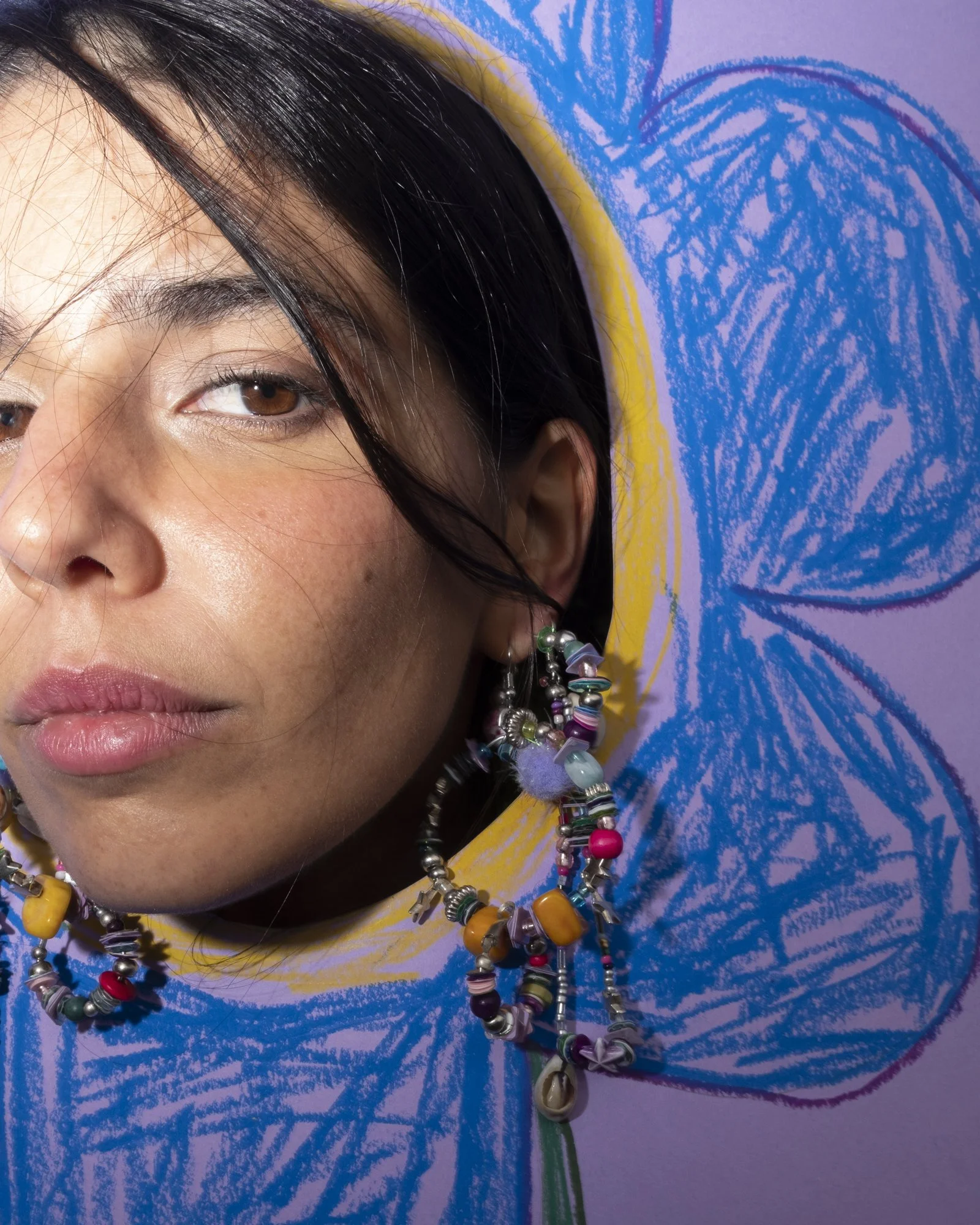 A woman with dark hair, wearing colorful bead earrings, stands in front of a blue and yellow floral artwork.