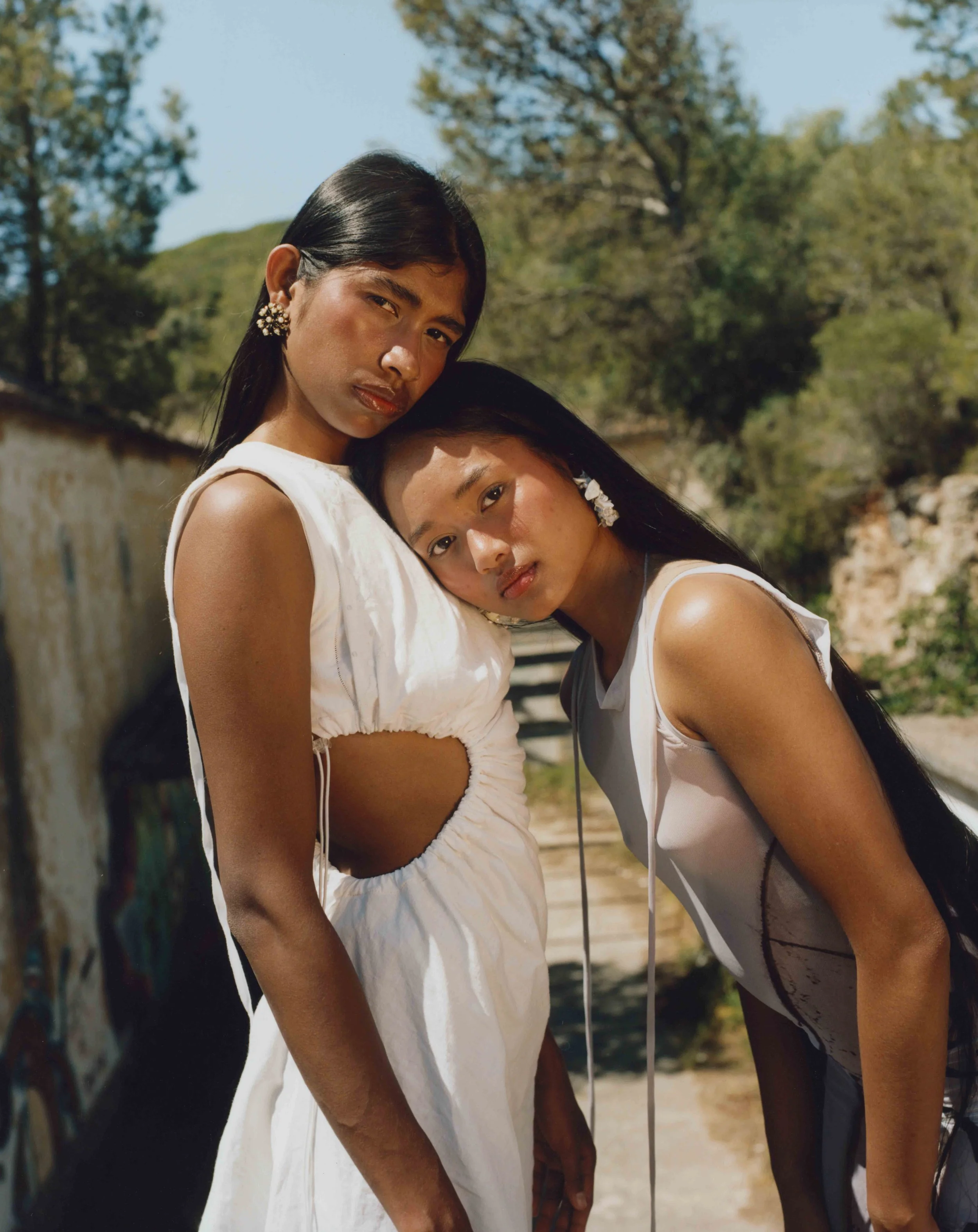 Two young women with long black hair and earrings stand close together outdoors during daytime, with one resting her head on the other's shoulder and both looking at the camera.
