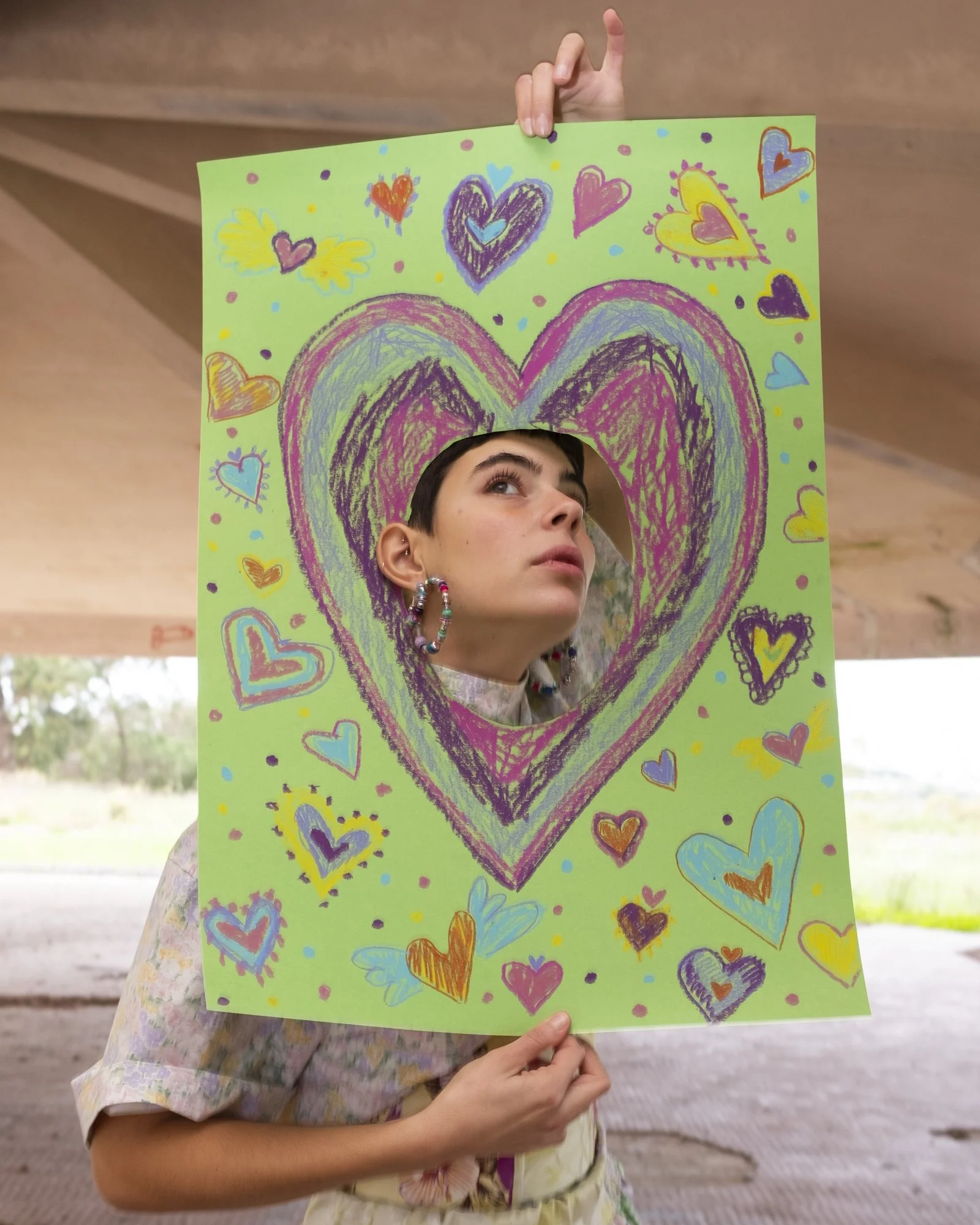 A person holds a colorful, hand-drawn heart-shaped mirror with a green background, surrounded by smaller hearts and doodles, reflecting their face with a playful expression.