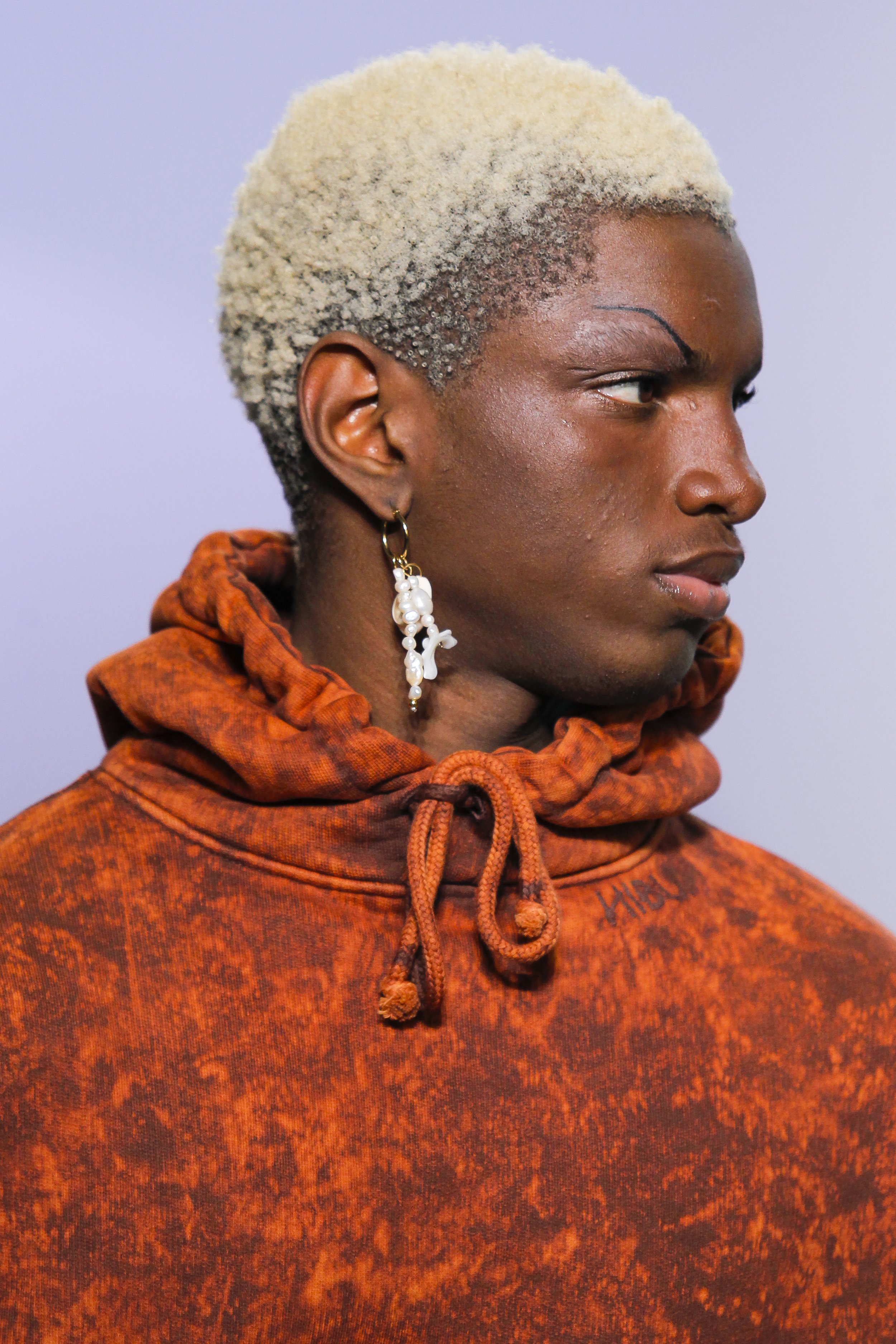 Profile of a person with short, bleached blonde hair, wearing a brown sweatshirt with an orange pattern and a pearl and gold earring, against a plain background.