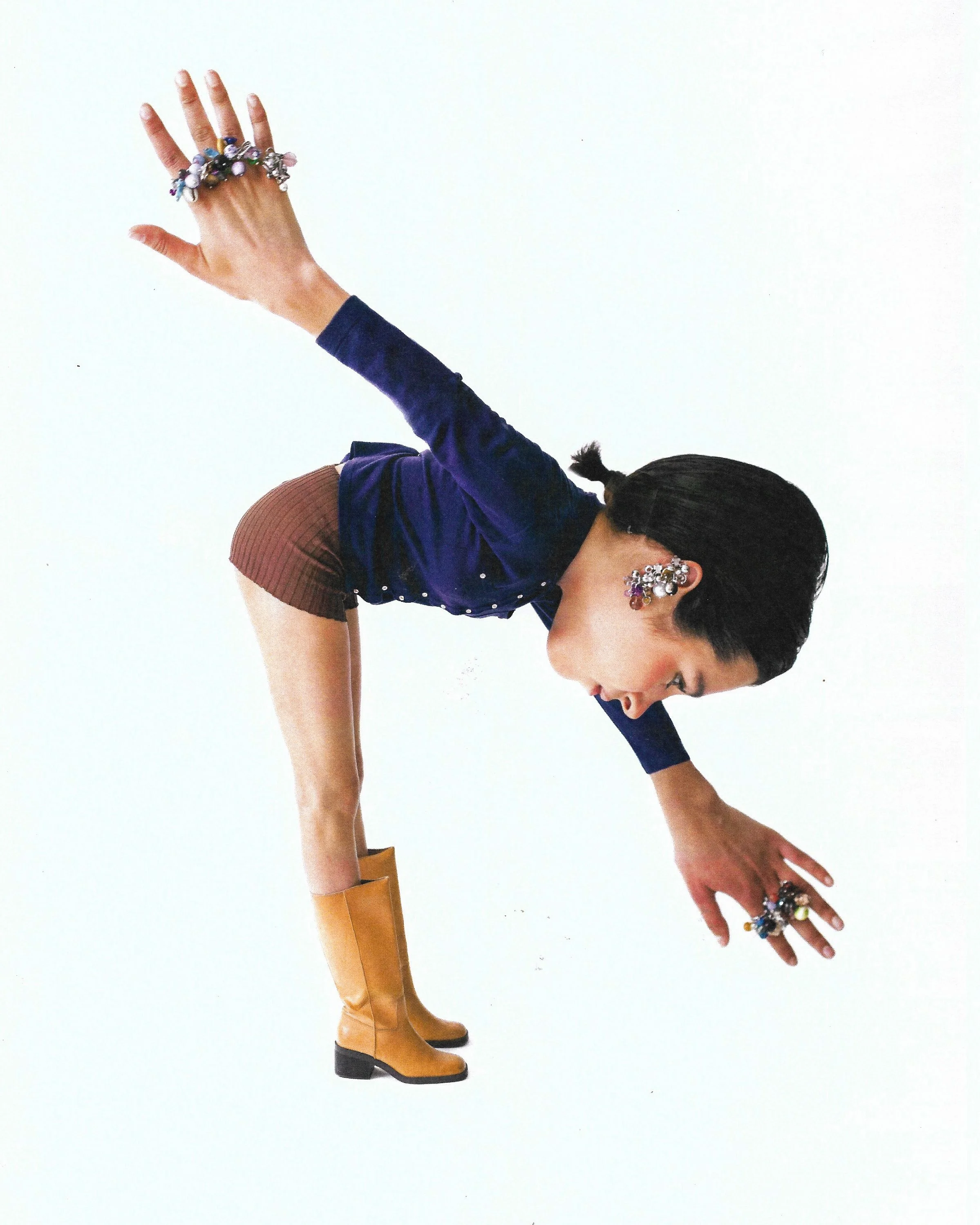 A woman dressed in a blue velvet jacket, brown skirt, and yellow boots performing a dance pose with one hand touching the ground and the other hand raised, adorned with jewelry and earrings, against a plain white background.