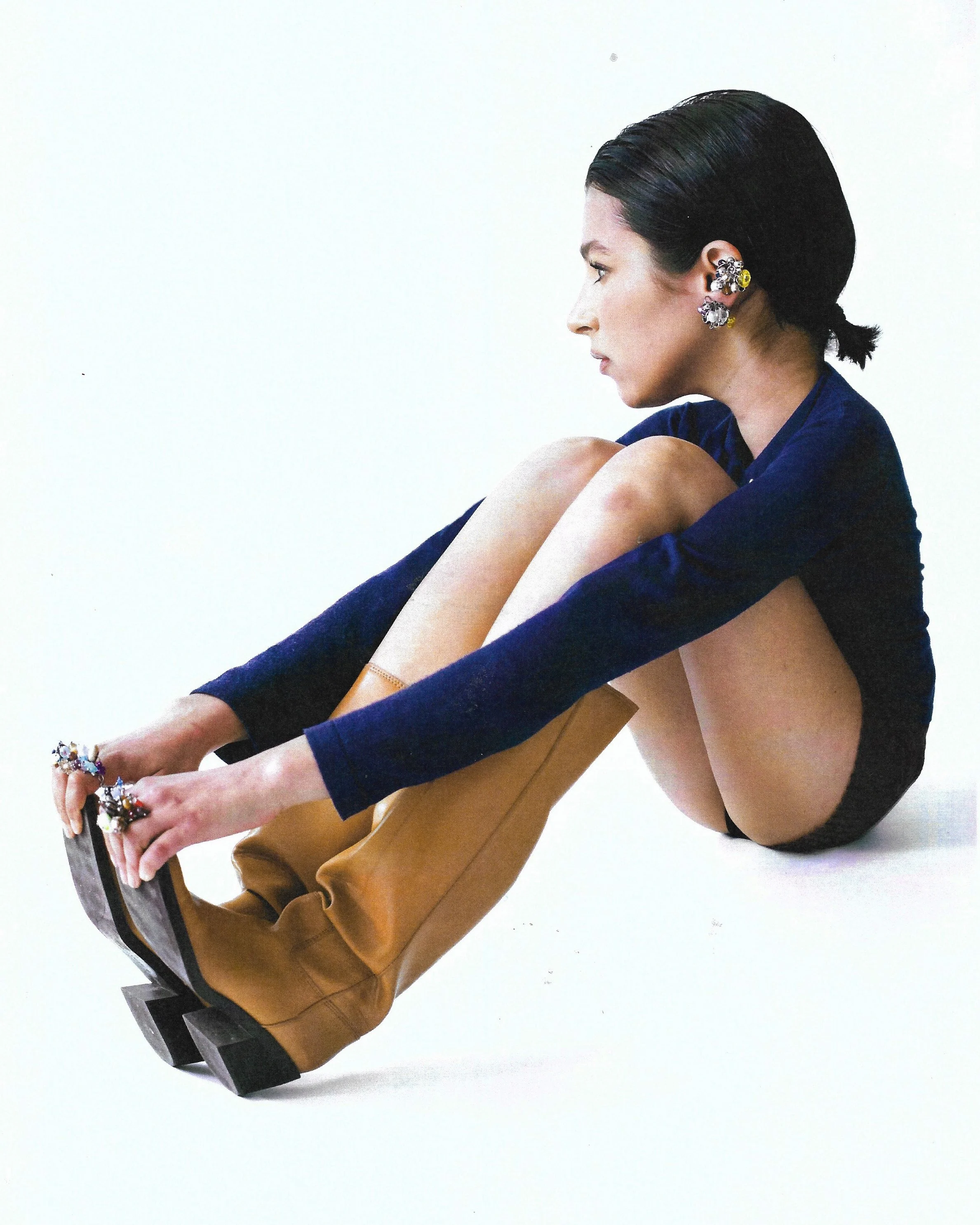 A woman sitting on the floor with her knees drawn up, wearing tan boots, a navy blue top, and black shorts, with her dark hair in a low ponytail and multiple earrings.