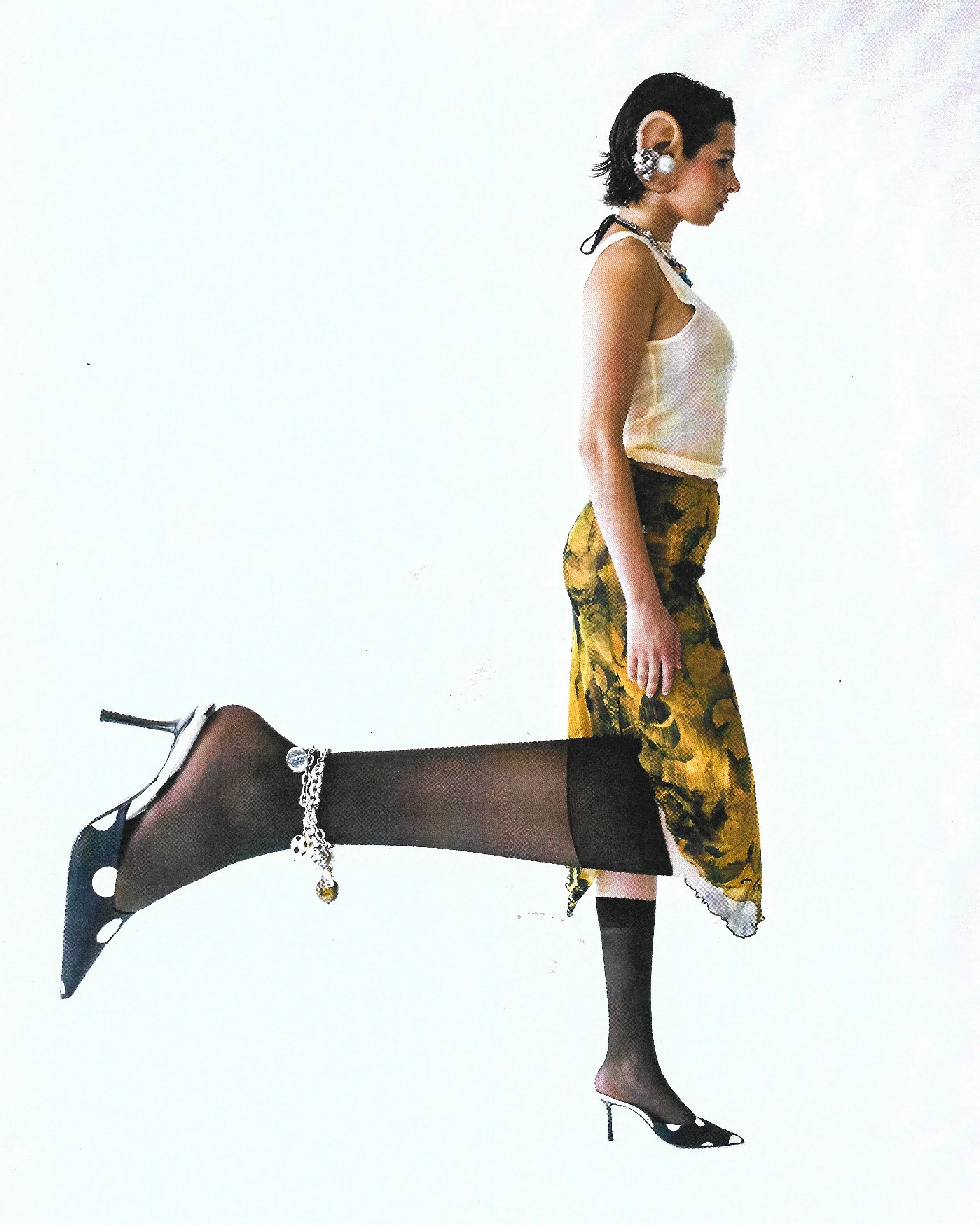 A woman with short black hair, wearing a white tank top, yellow and black patterned skirt, black sheer stockings with an anklet, and polka-dot high heels, stands against a plain white background.