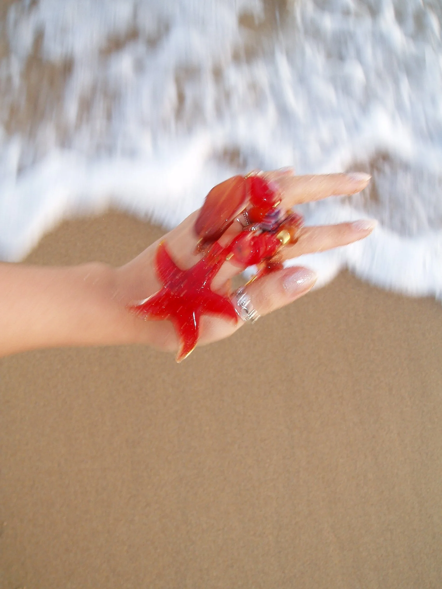 Person holding a small red starfish on the beach with sand and ocean waves in the background.
