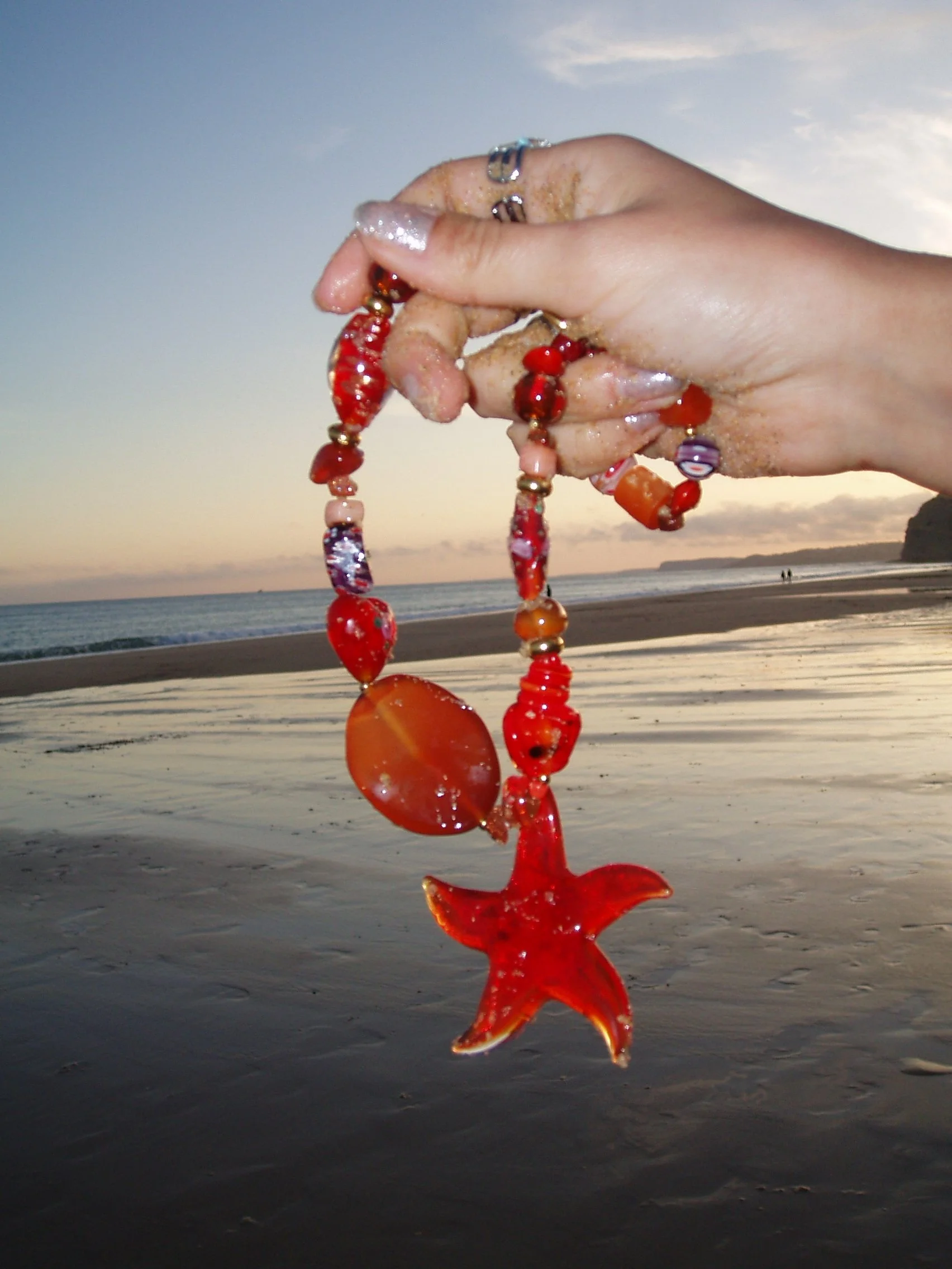 A hand holding a red and orange starfish-shaped shell necklace against a beach at sunset with sand, the ocean, and a cloudy sky in the background.