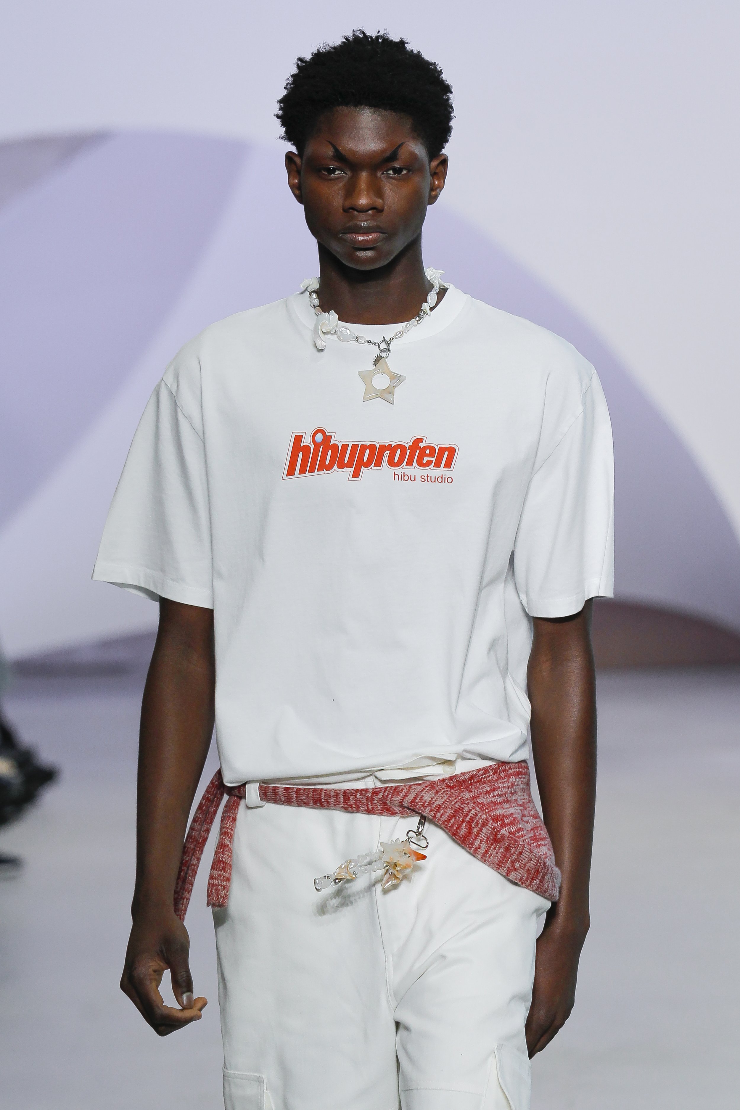 A male model walking on a runway wearing a white t-shirt with red text and a red knitted sweater tied around his waist, paired with white cargo pants. The model has short, curly black hair and is wearing a necklace with star and shell charms.