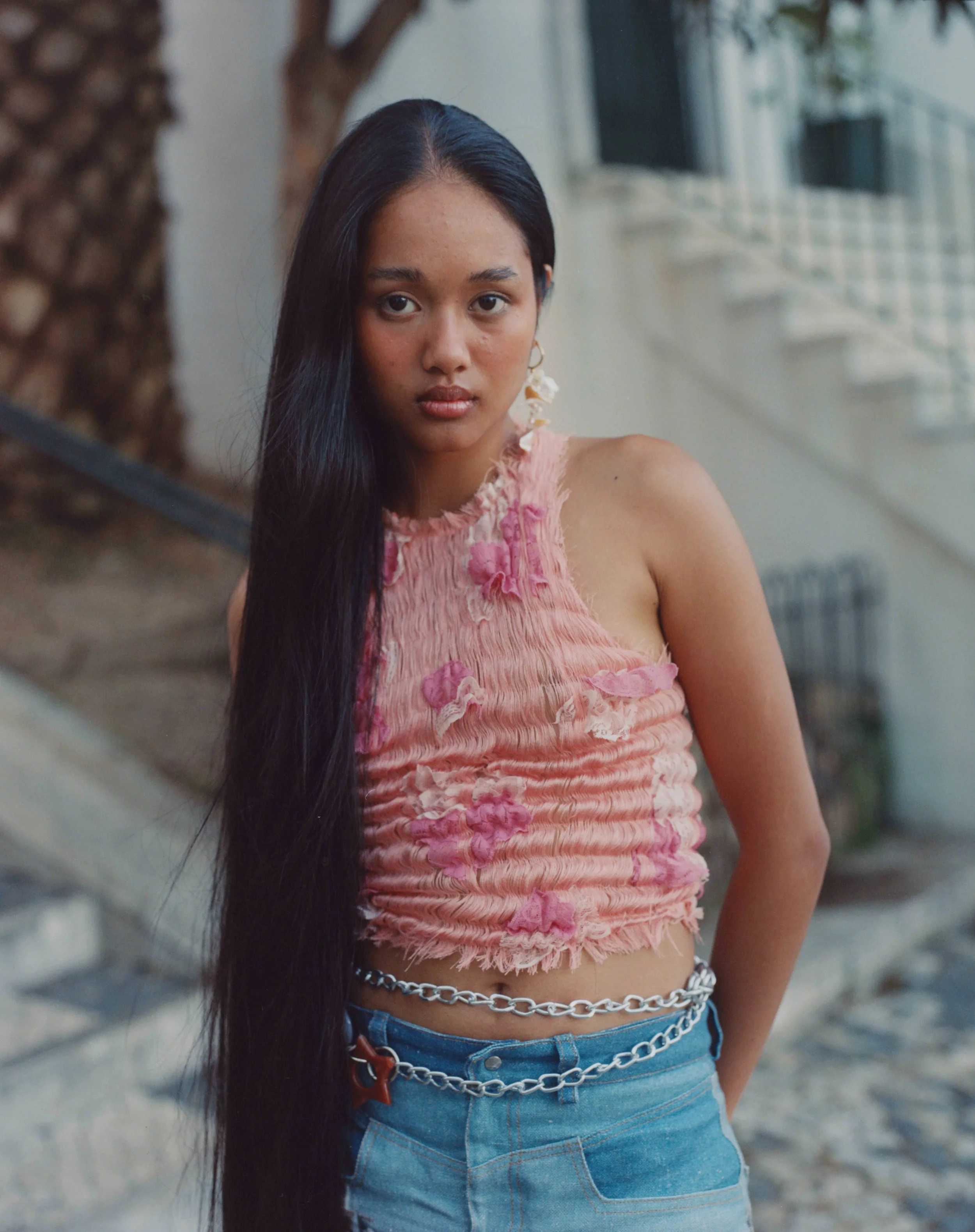 Young woman with long black hair wearing a pink floral sleeveless top and blue jeans with chains, standing outdoors near a tree and a building.
