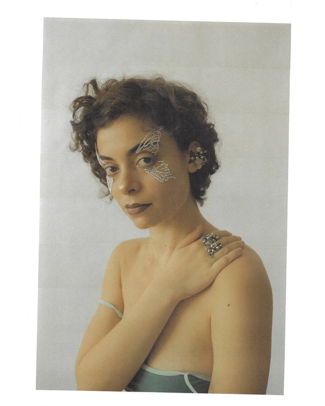 Portrait of a woman with short curly hair wearing rhinestone face decorations resembling butterfly wings, reaching her collarbone, and silver rings on her fingers, against a plain white background.