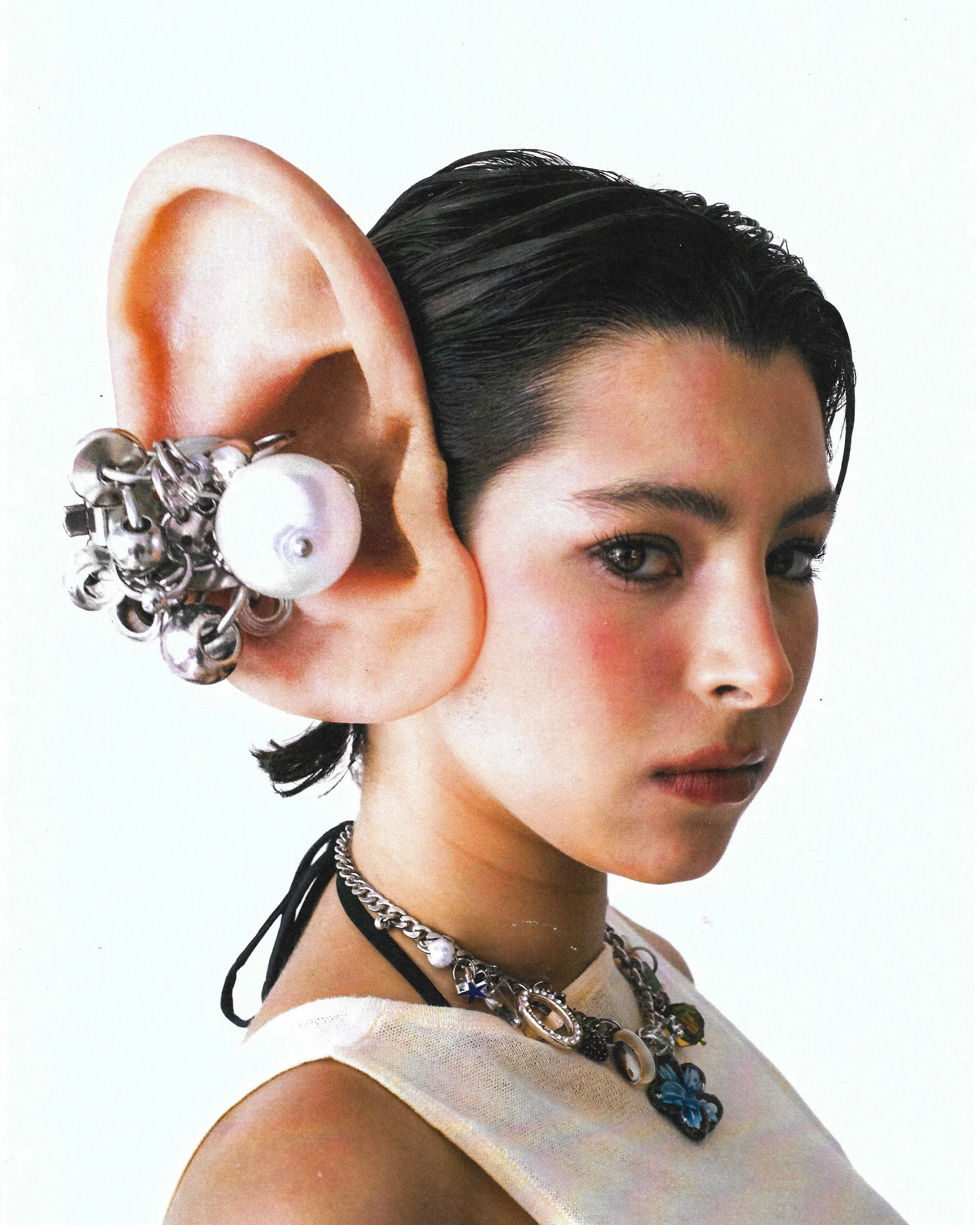 Close-up of a woman's face with short dark hair, wearing layered necklaces with charms, earrings, and a white top. There is an oversized ear prop with jewelry attached to her left ear.