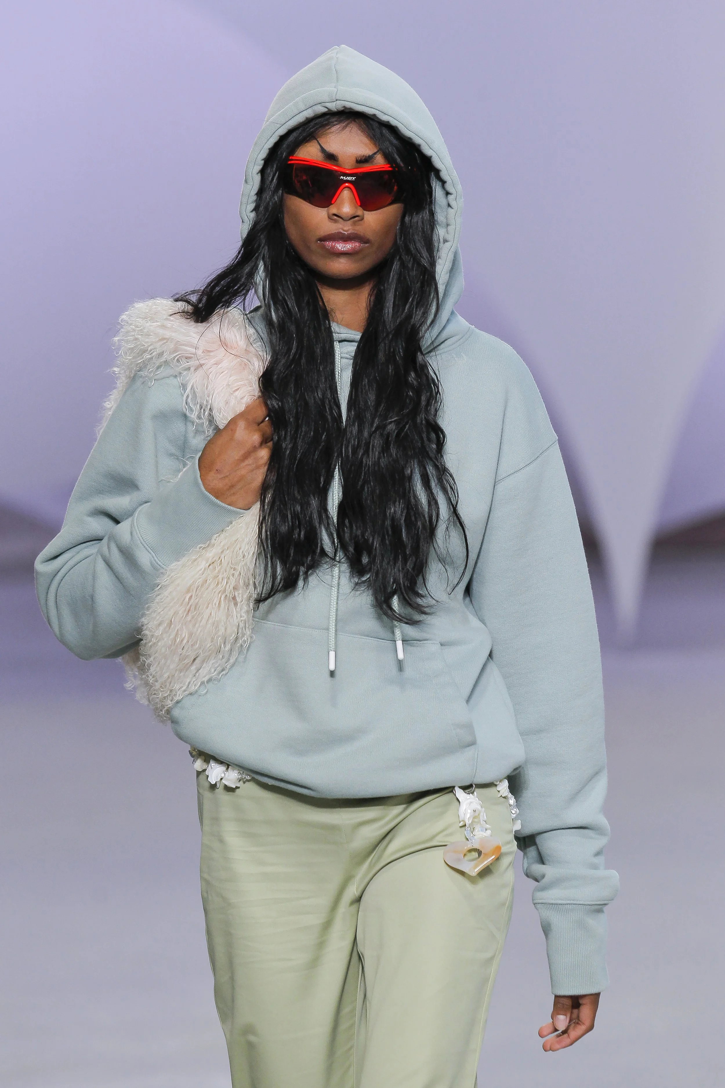 A woman with long black wavy hair wearing a light blue hoodie, red sunglasses, and beige pants walking on a runway at a fashion show.